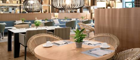 A modern restaurant with round tables, wicker chairs, and hanging pendant lights.