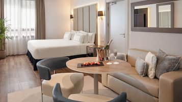 A hotel room with a bed, a sofa, a round table with a bottle and glasses, and a potted plant.