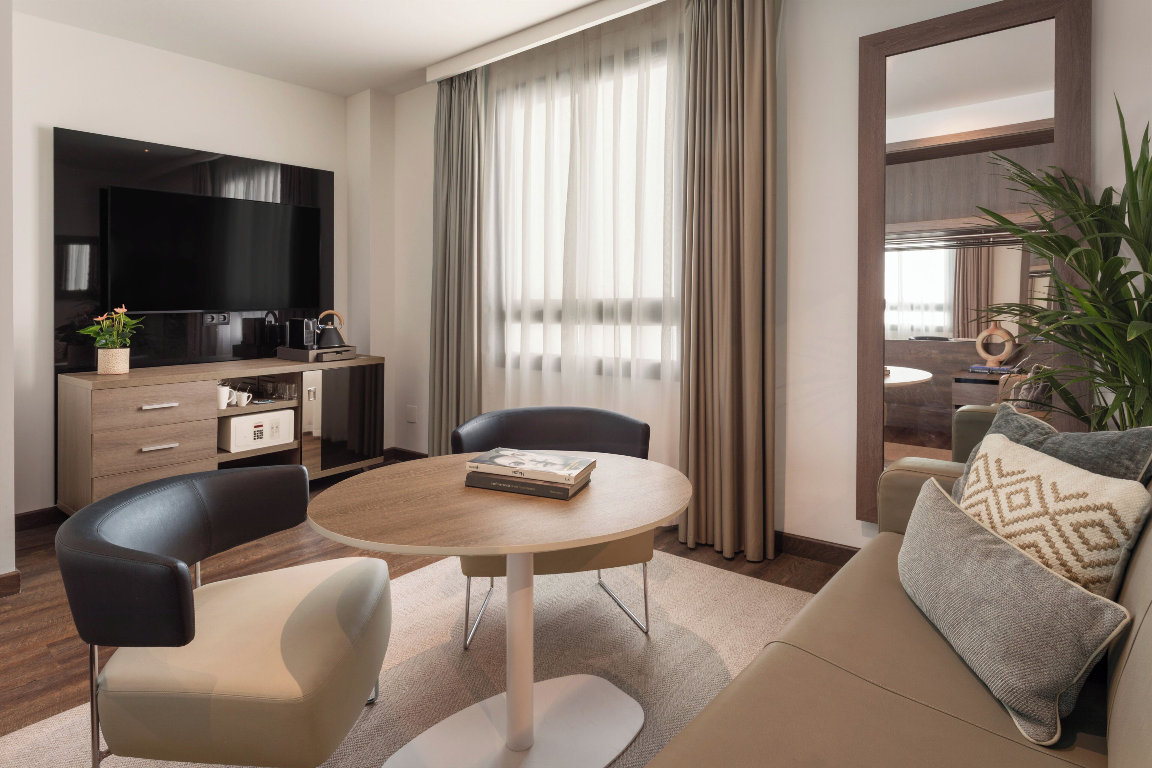 Habitación de hotel moderna con una televisión de pantalla plana, una mesa de comedor redonda, dos sillas, un sofá, una cafetera, una planta y un gran ventanal con cortinas.