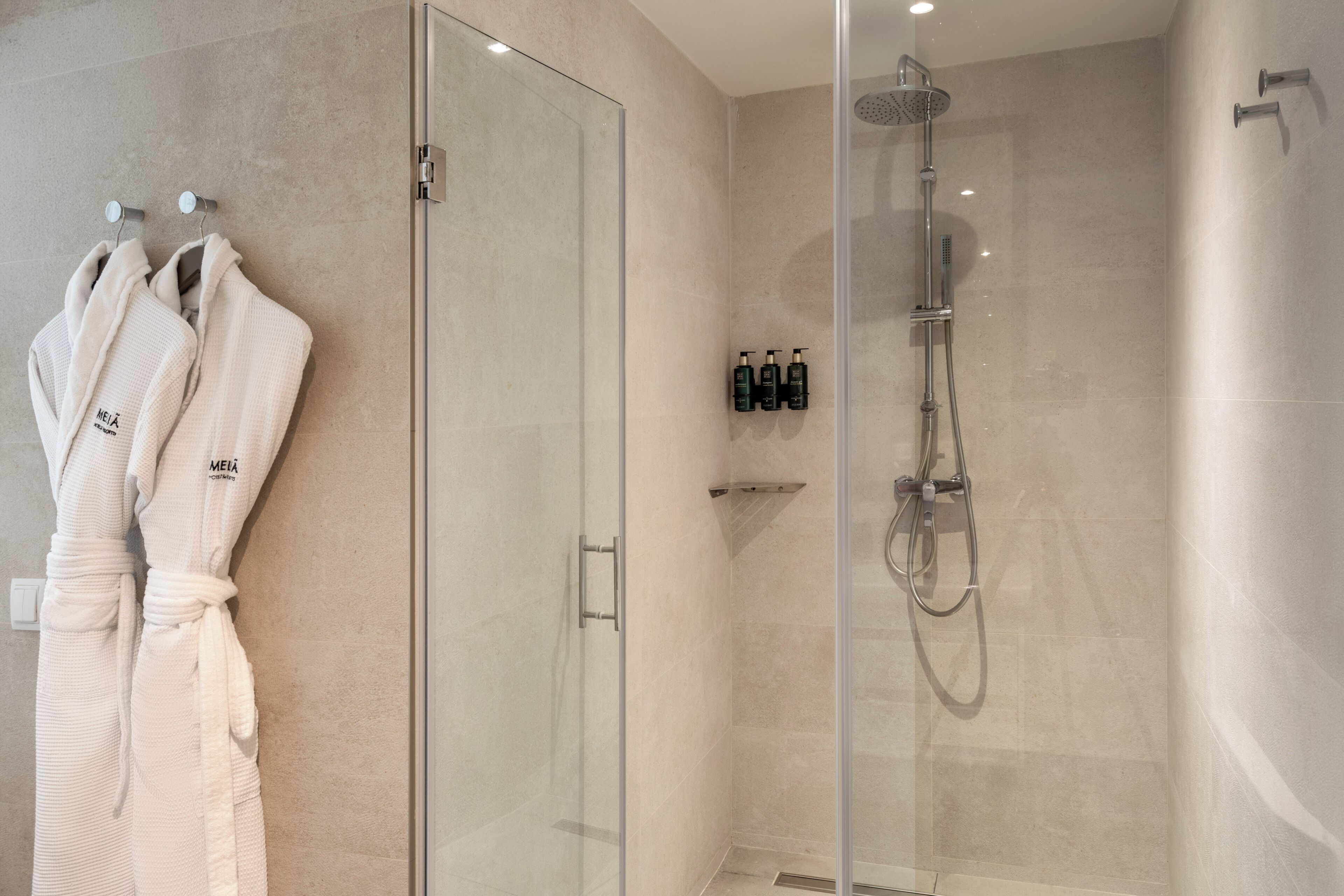 A modern bathroom with a glass shower enclosure, a glass door, and two white bathrobes hanging on hooks.