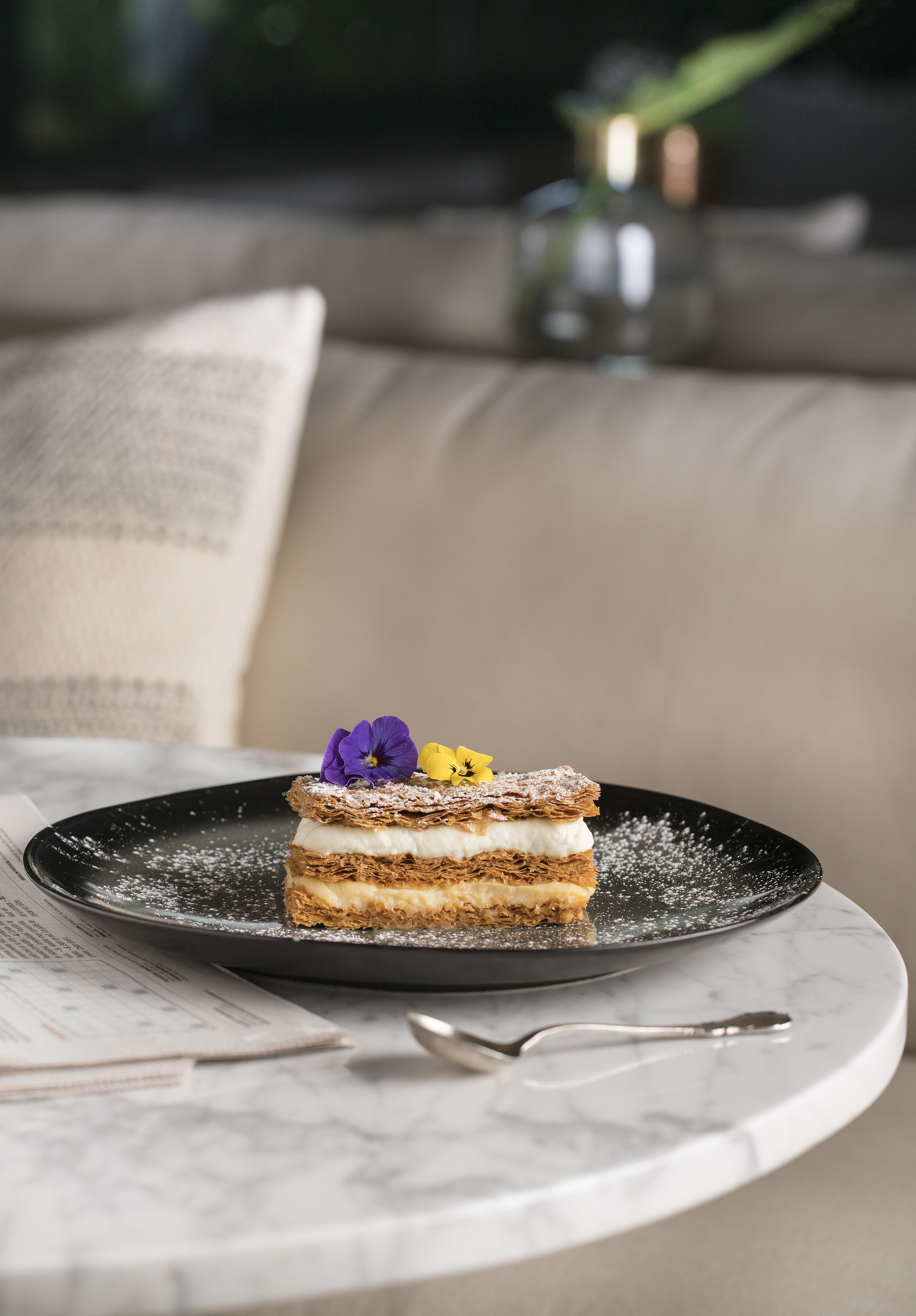 A slice of layered cake on a black plate with powdered sugar, placed on a marble table.
