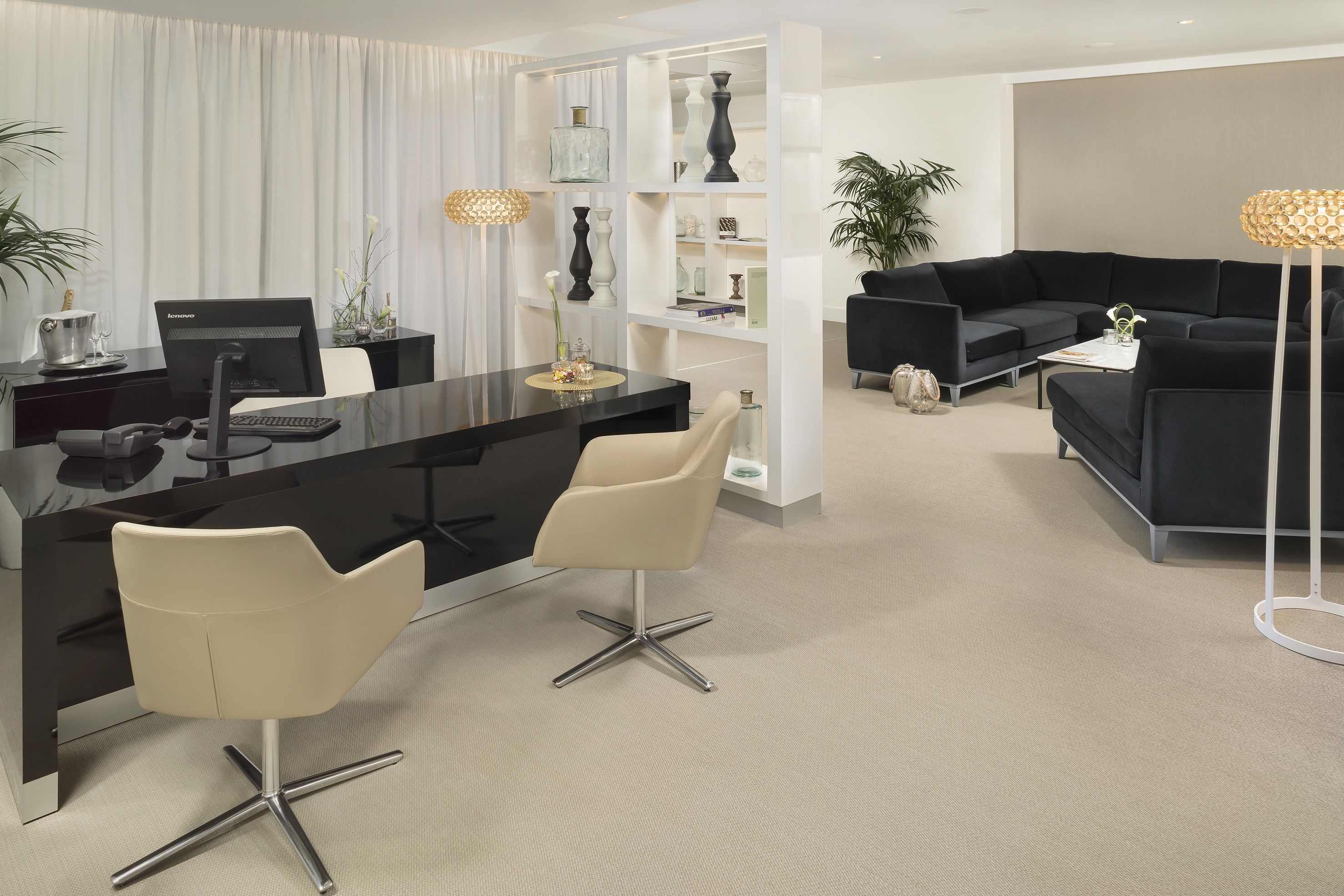 A modern living room with a black sofa, a black coffee table, and a beige chair. There is a desk with a computer and a lamp.