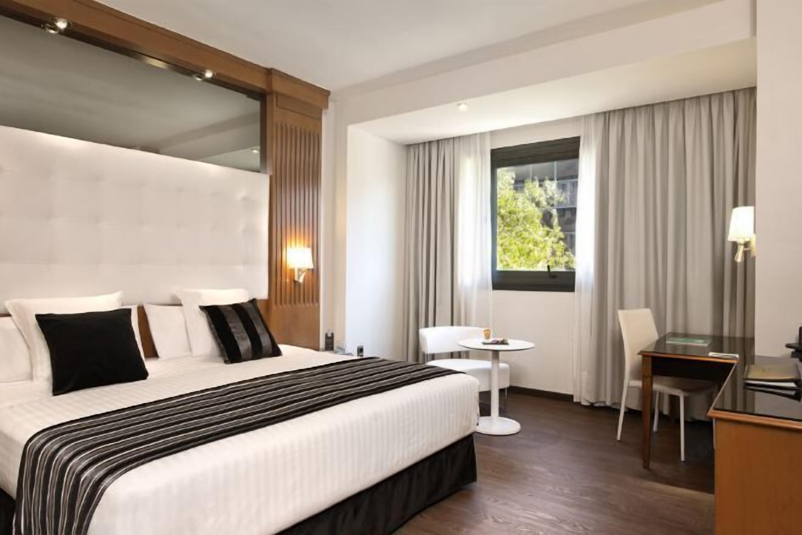A hotel room with a large bed, a desk, a chair, and a window with curtains.