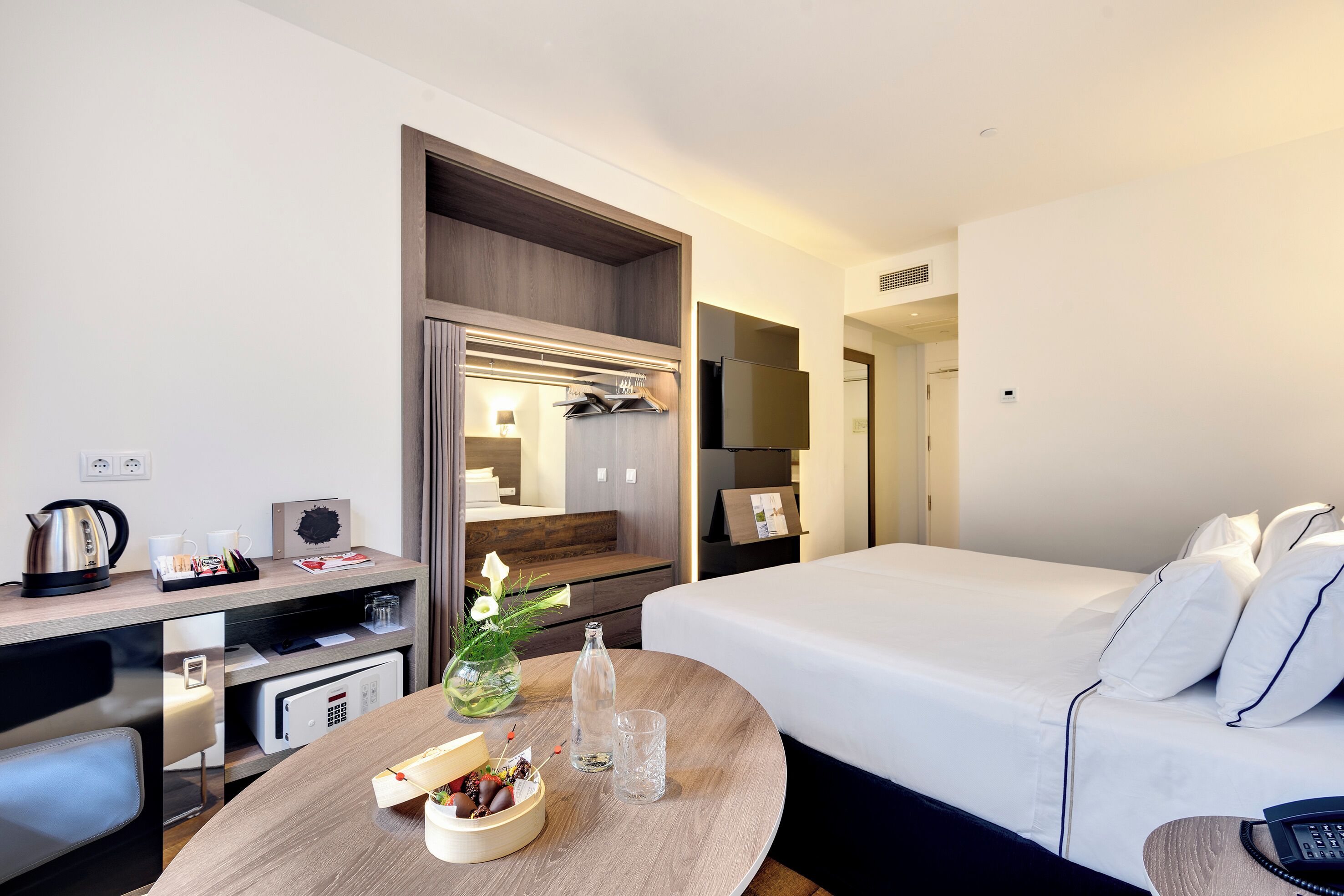 A modern hotel room with a neatly made bed, a desk with a kettle, a microwave, and a small table with a vase and a bottle.