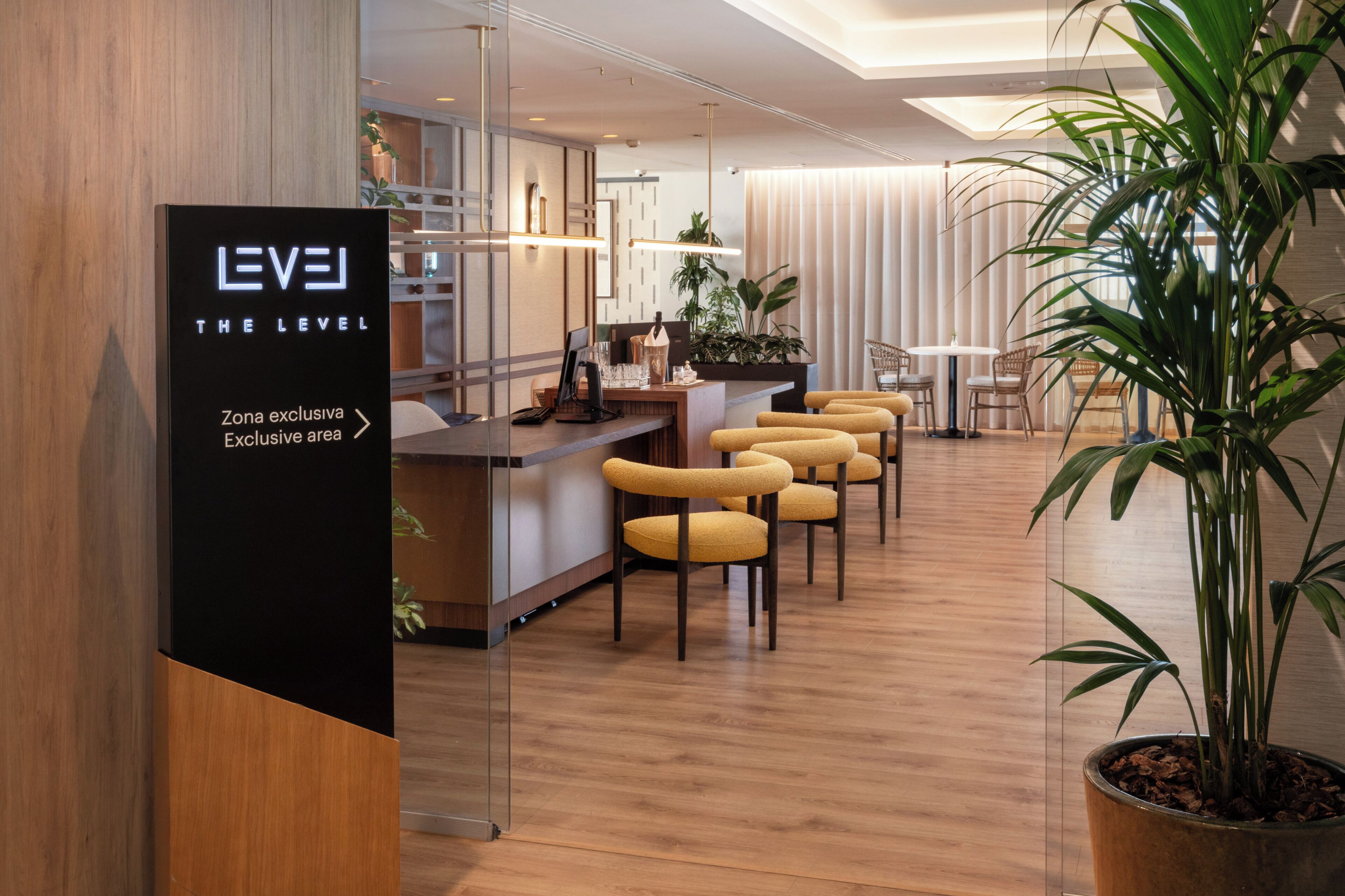 A modern hotel lobby with a reception desk, bar area, yellow chairs, potted plants, and a sign indicating an exclusive area.