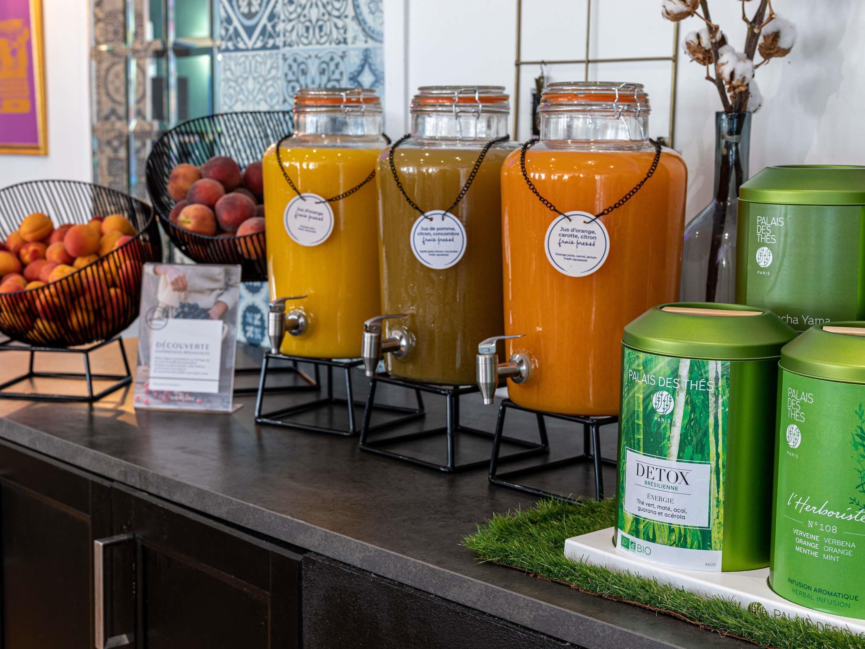 A counter with large glass containers of orange juice, a bowl of fruit, and green herbal infusion bottles.