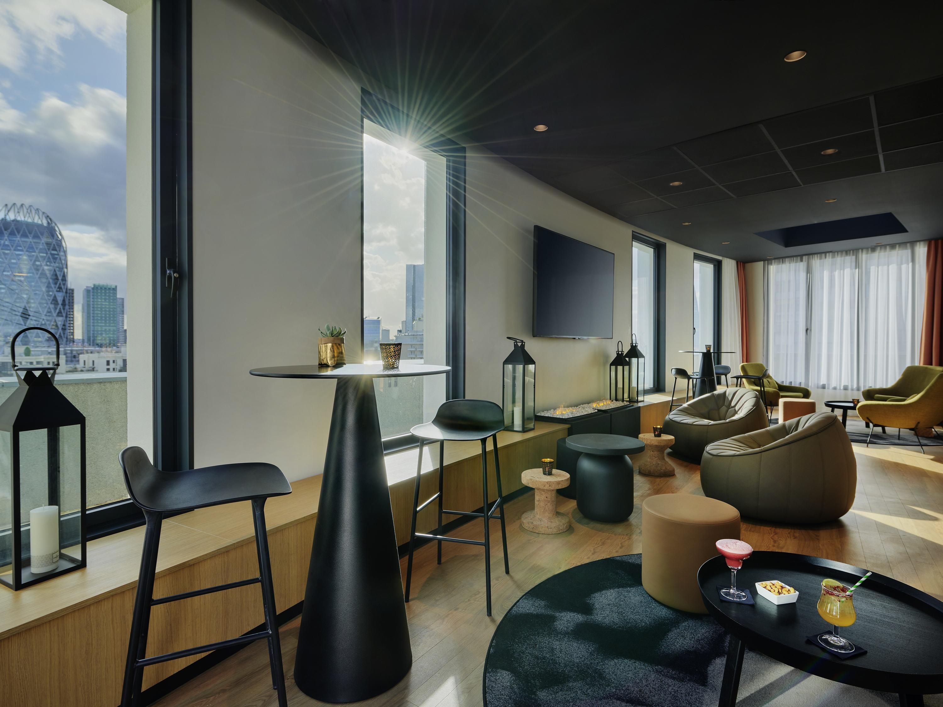 A modern hotel room with a city view, a round table, bar stools, a sofa, and a flat-screen TV.