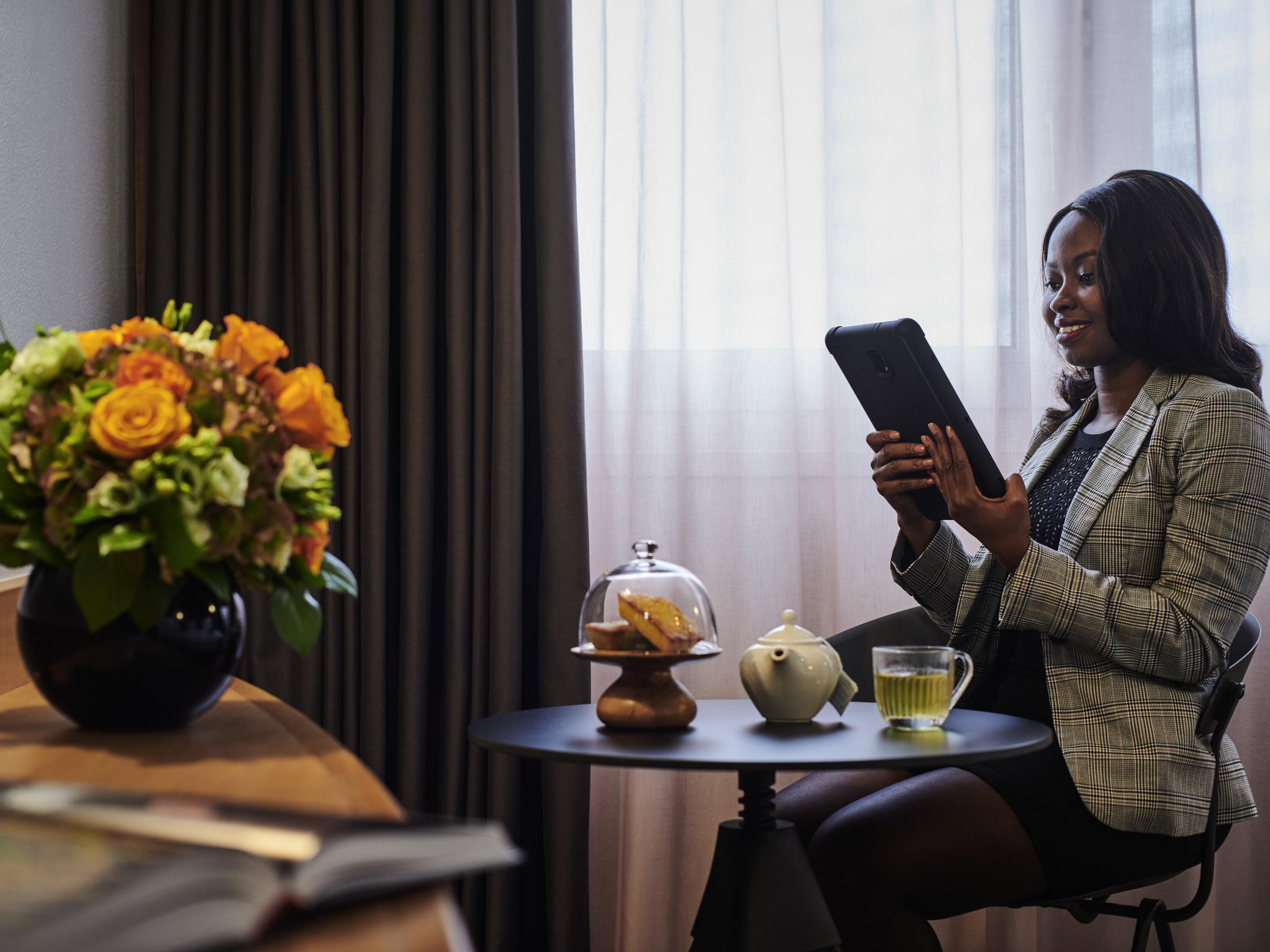 A person sitting at a table with a smartphone, a vase of flowers, a teapot, and a glass of tea.