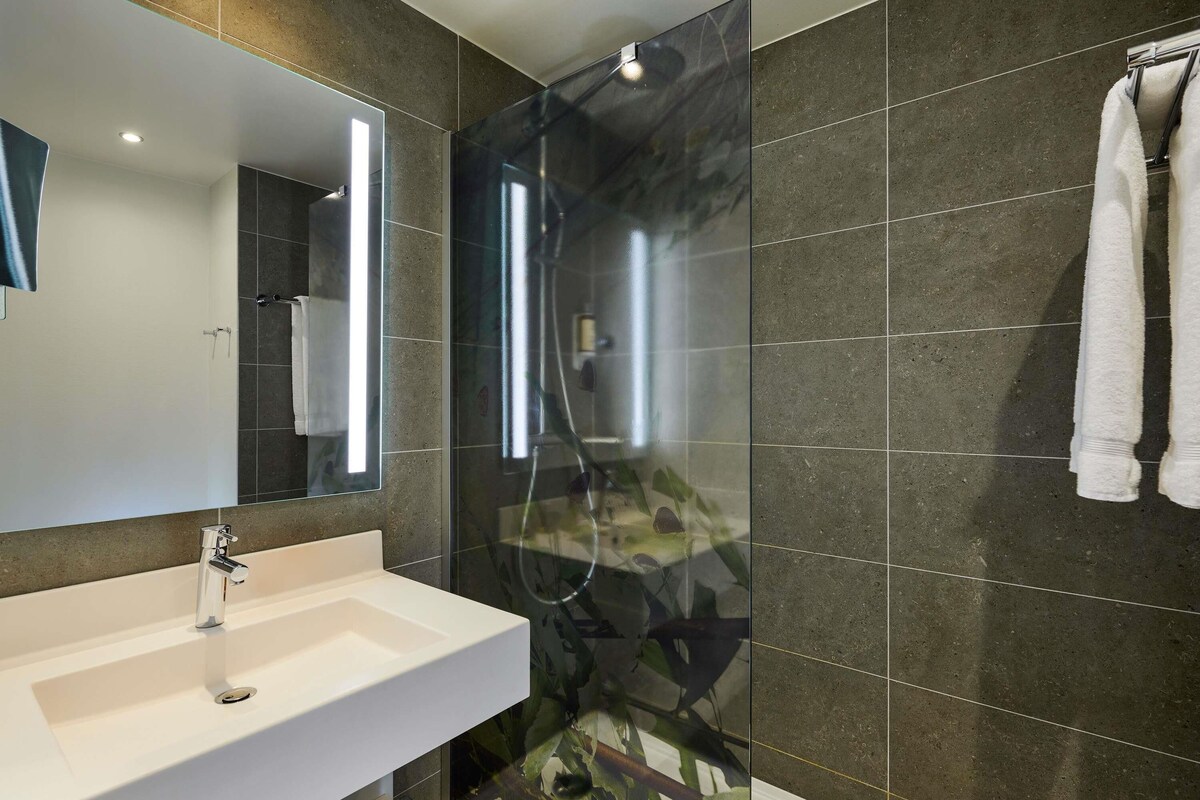 A bathroom with a white sink, a mirror, a shower with a glass door, and dark tiled walls.