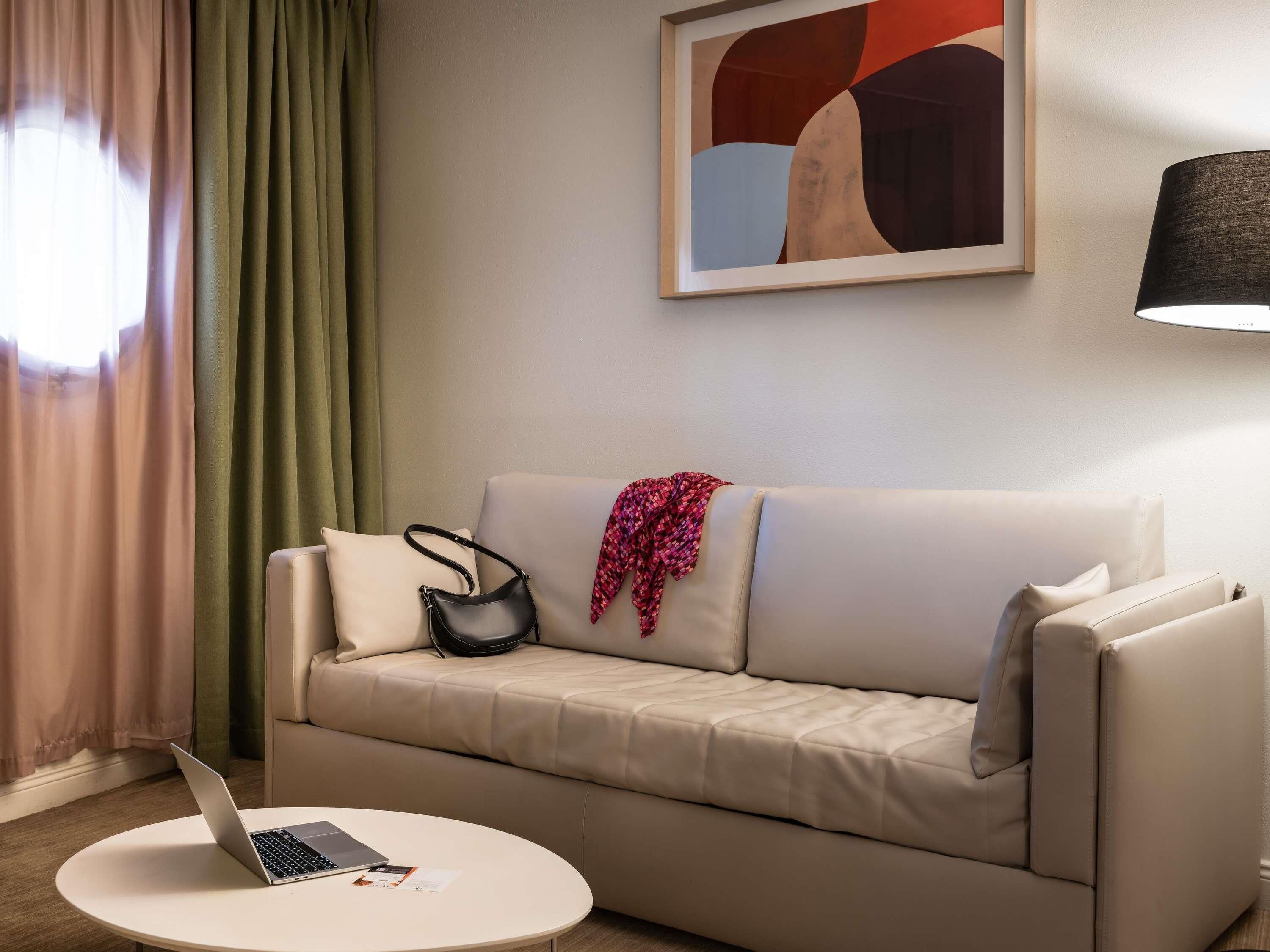 A modern living room with a beige sofa, a round coffee table, a laptop, a black handbag, and a colorful throw.