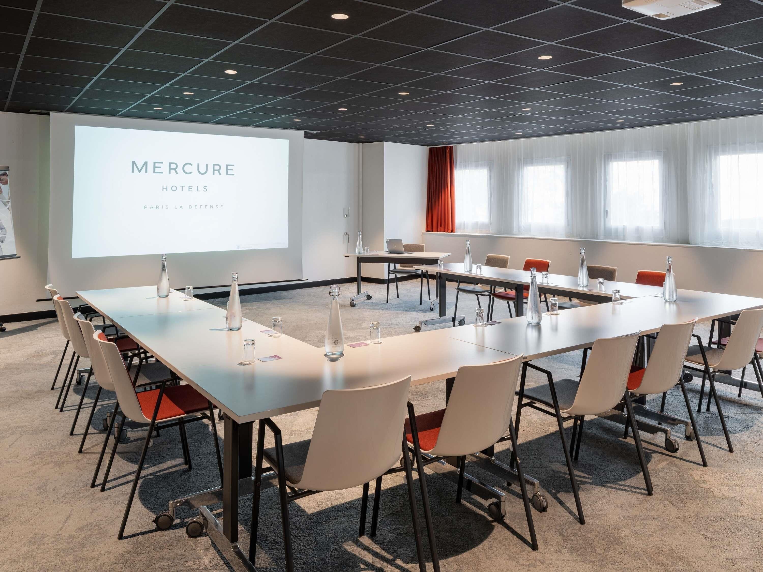 A conference room with a long table, chairs, and a projector screen displaying 'Mercure Hotels Paris La Défense'.