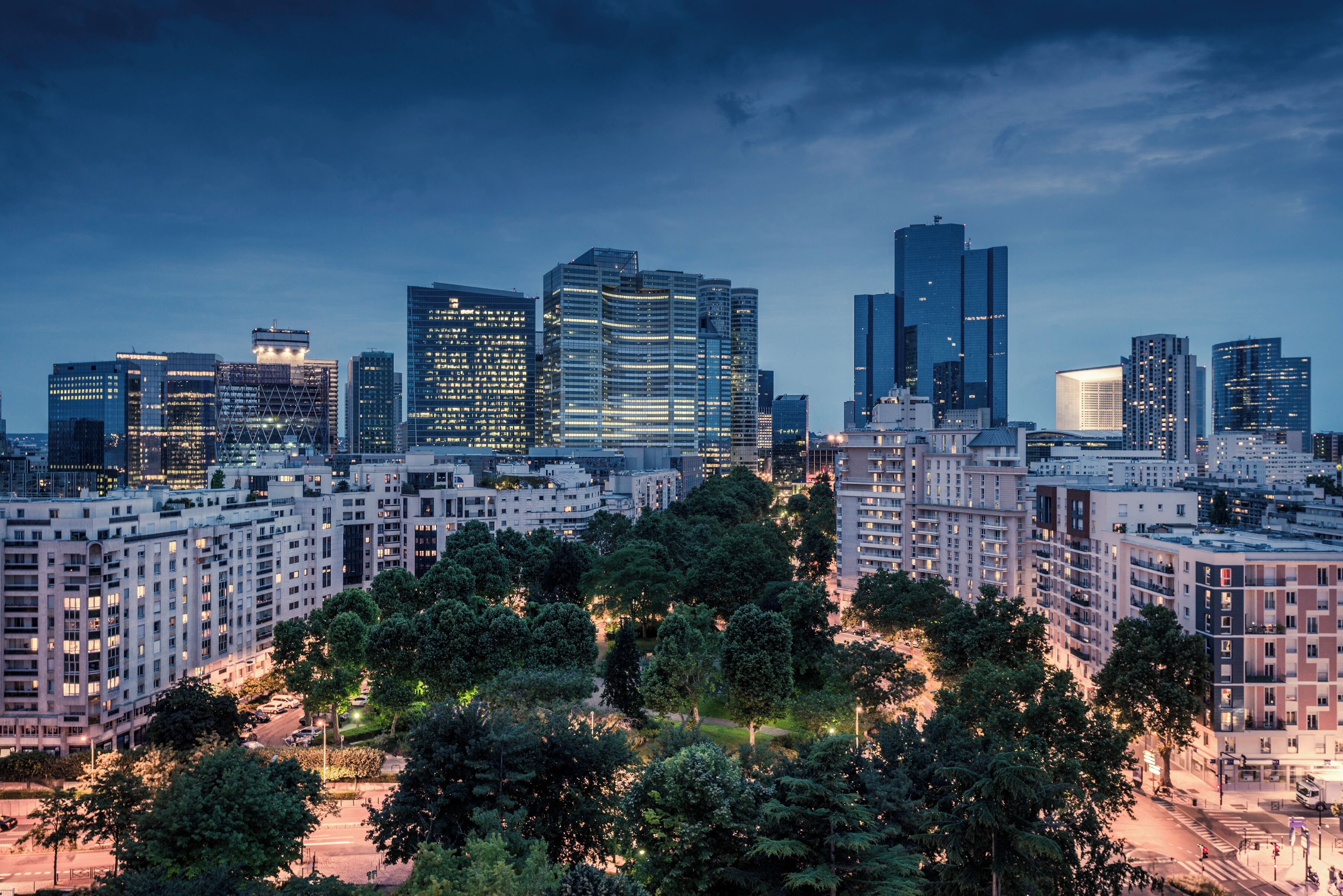 Un paysage urbain avec des immeubles modernes de grande hauteur, un espace vert arboré et un environnement urbain bien éclairé.