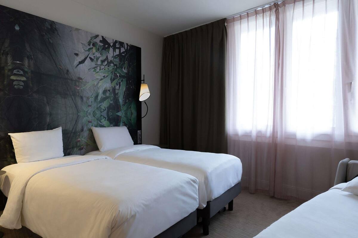 A hotel room with two beds, a large abstract painting on the wall, a lamp, and a window with sheer curtains.