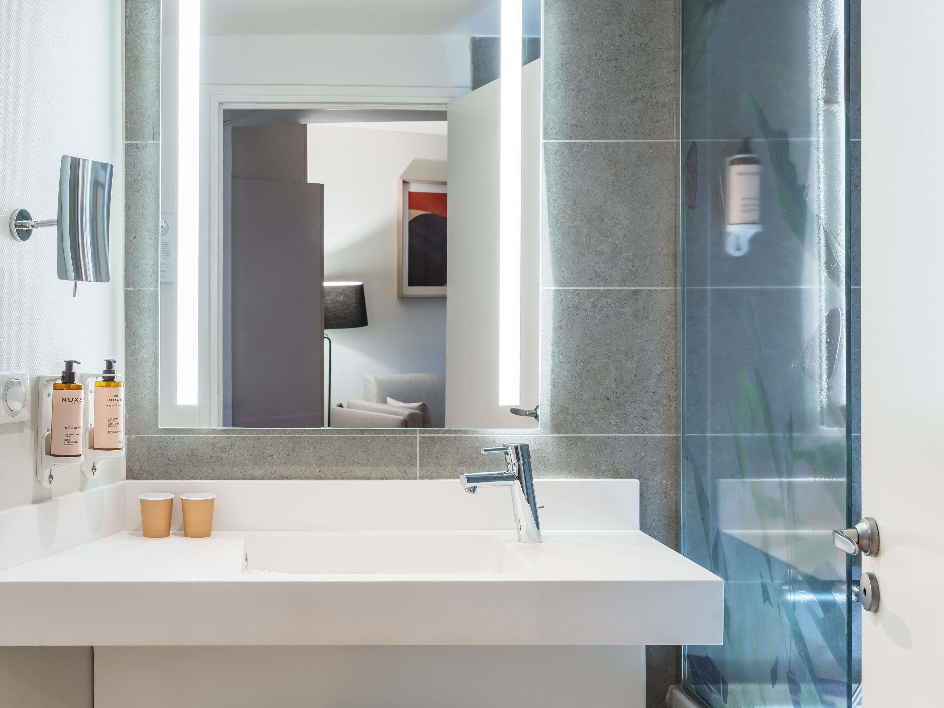 A modern bathroom with a white sink, a glass shower enclosure, and a mirror reflecting a bedroom.