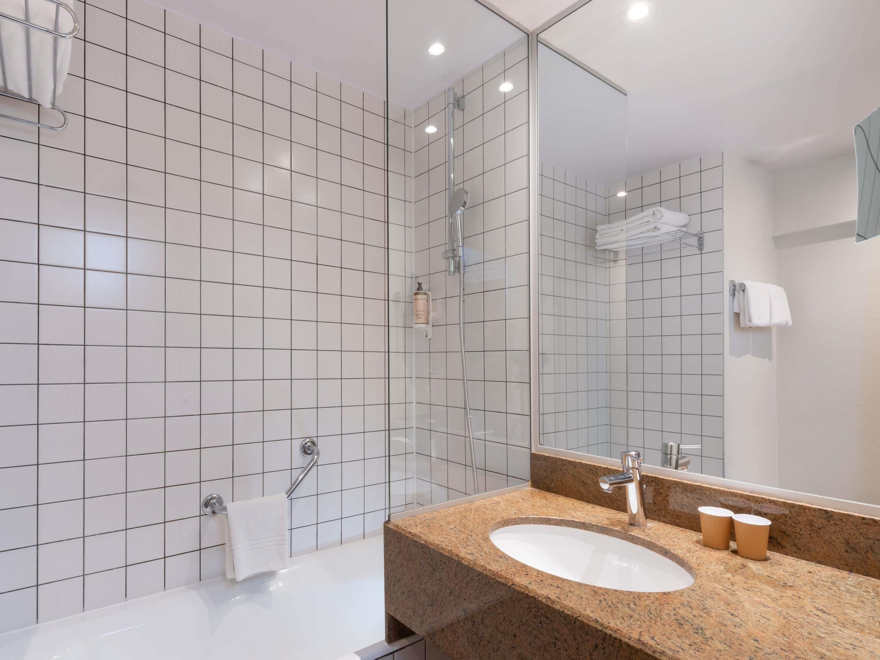 Een moderne badkamer met witte betegelde wanden, een grote spiegel, een wastafel met een granieten aanrechtblad en een douchehoek.