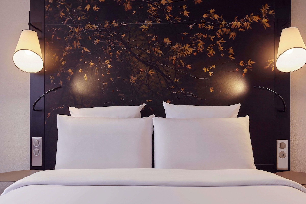 A hotel room with a bed, two bedside lamps, and a wall-mounted artwork.