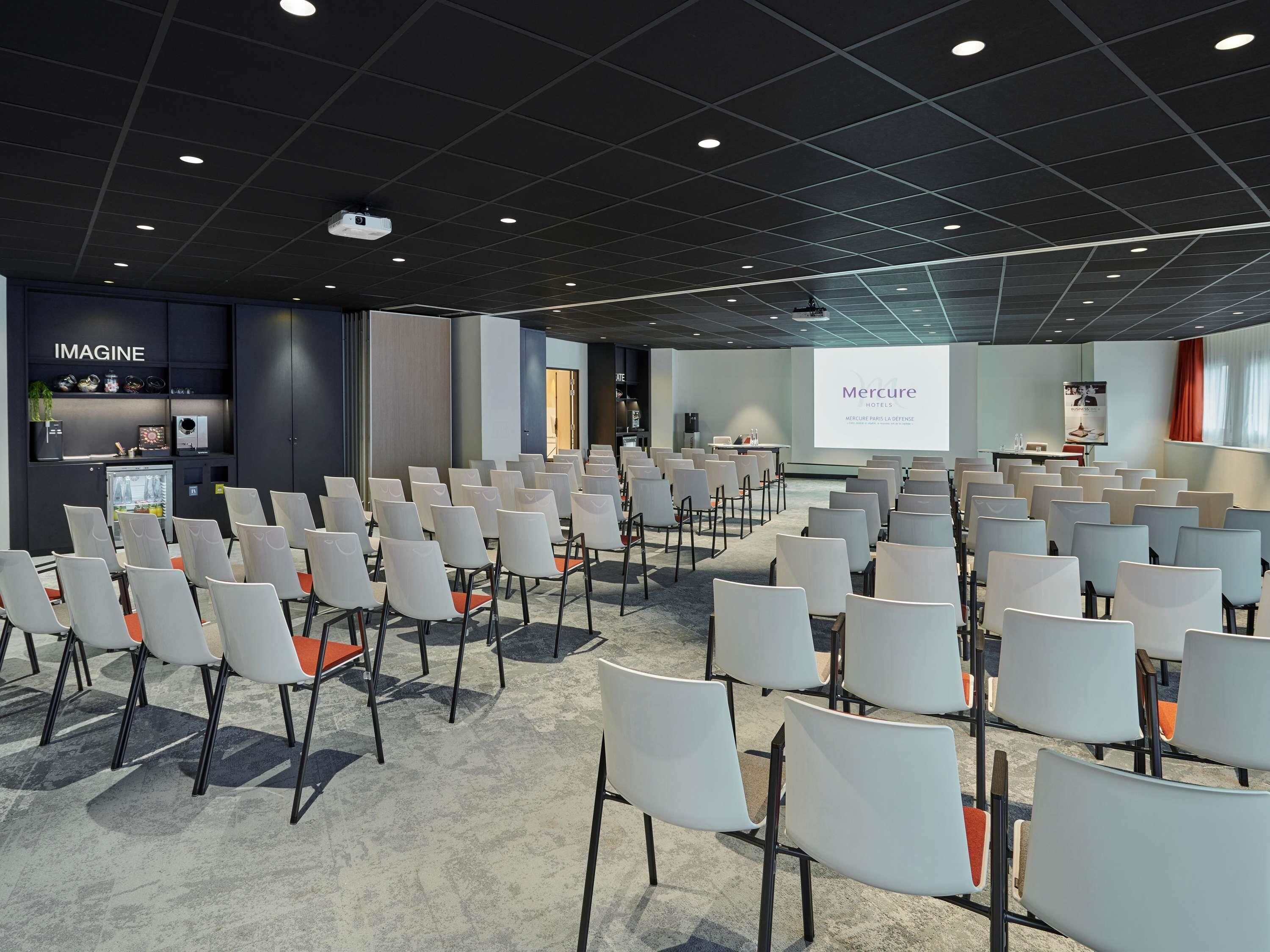 Un salón de conferencias con filas de sillas, una pantalla de proyector y una zona para presentaciones.