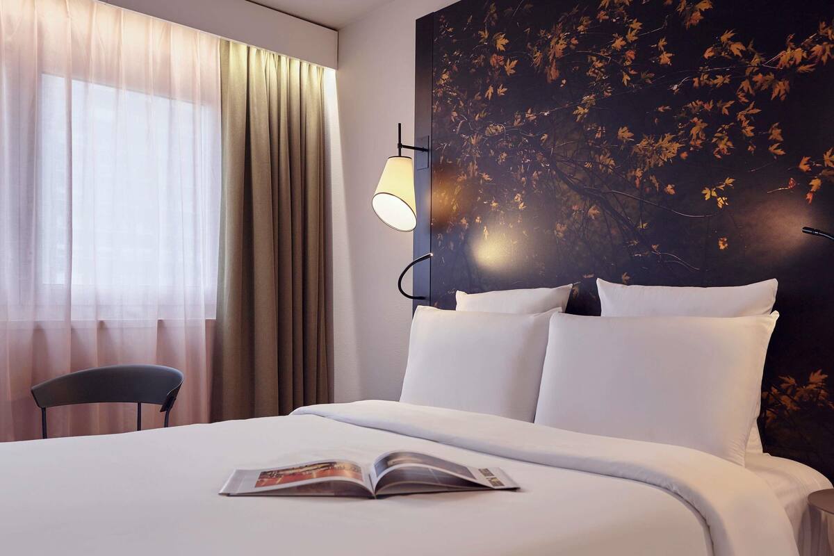 A hotel room with a bed, a chair, a lamp, and a wall with a tree mural.