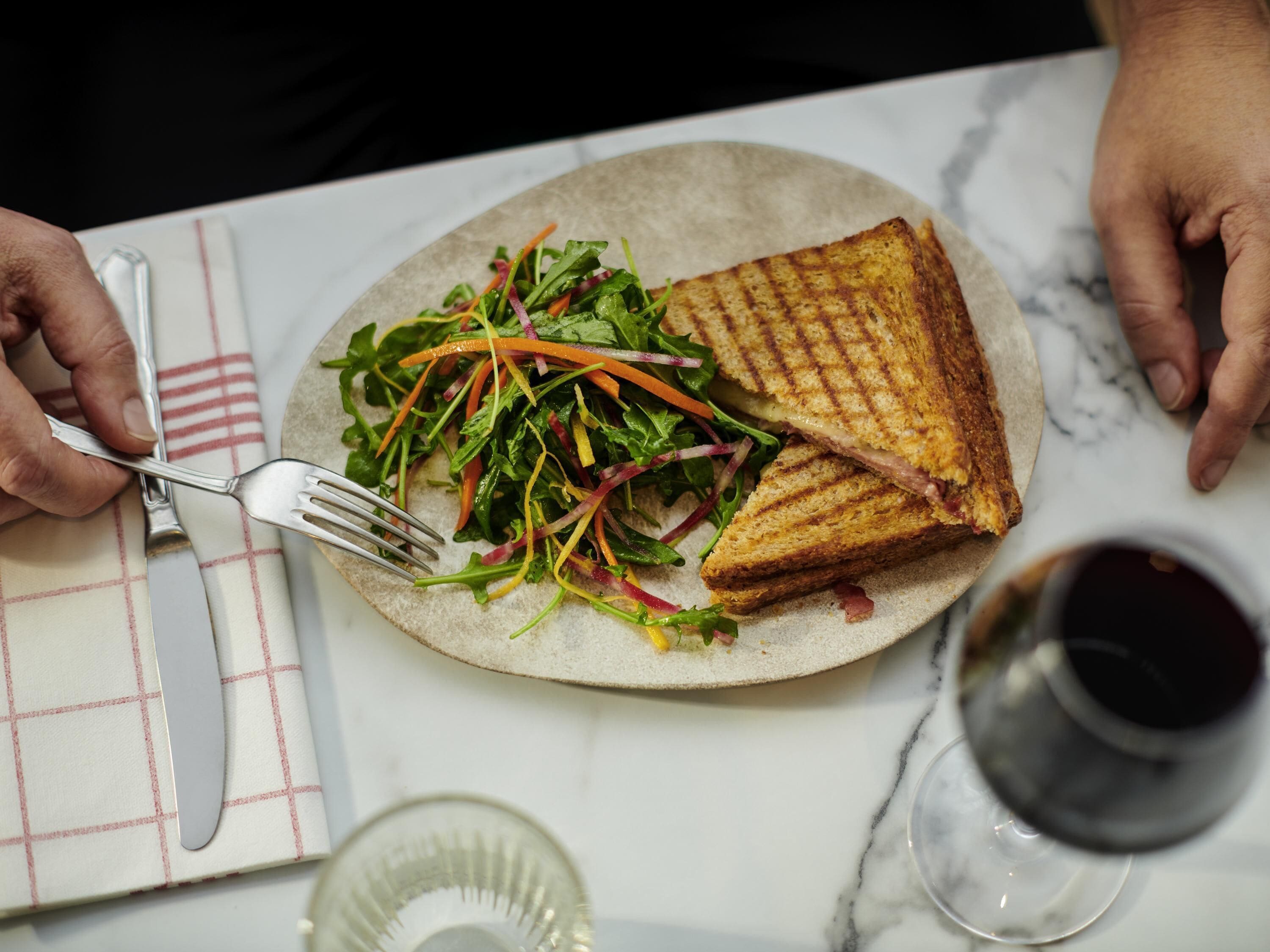 Um prato com sanduíche grelhado e salada, um garfo, uma faca e um copo de vinho tinto sobre uma mesa de mármore.