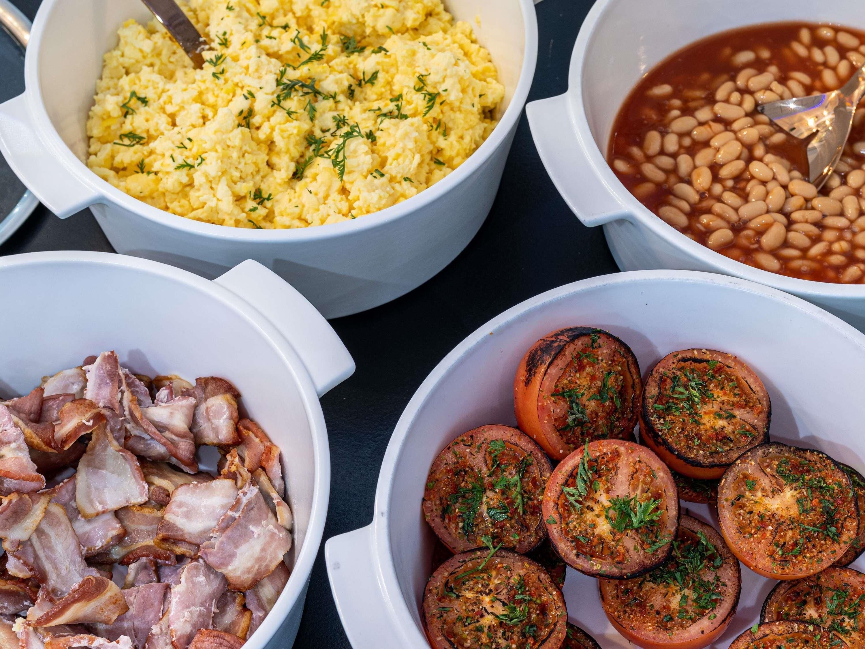 A table with four white bowls containing scrambled eggs, baked beans, bacon, and roasted tomatoes.