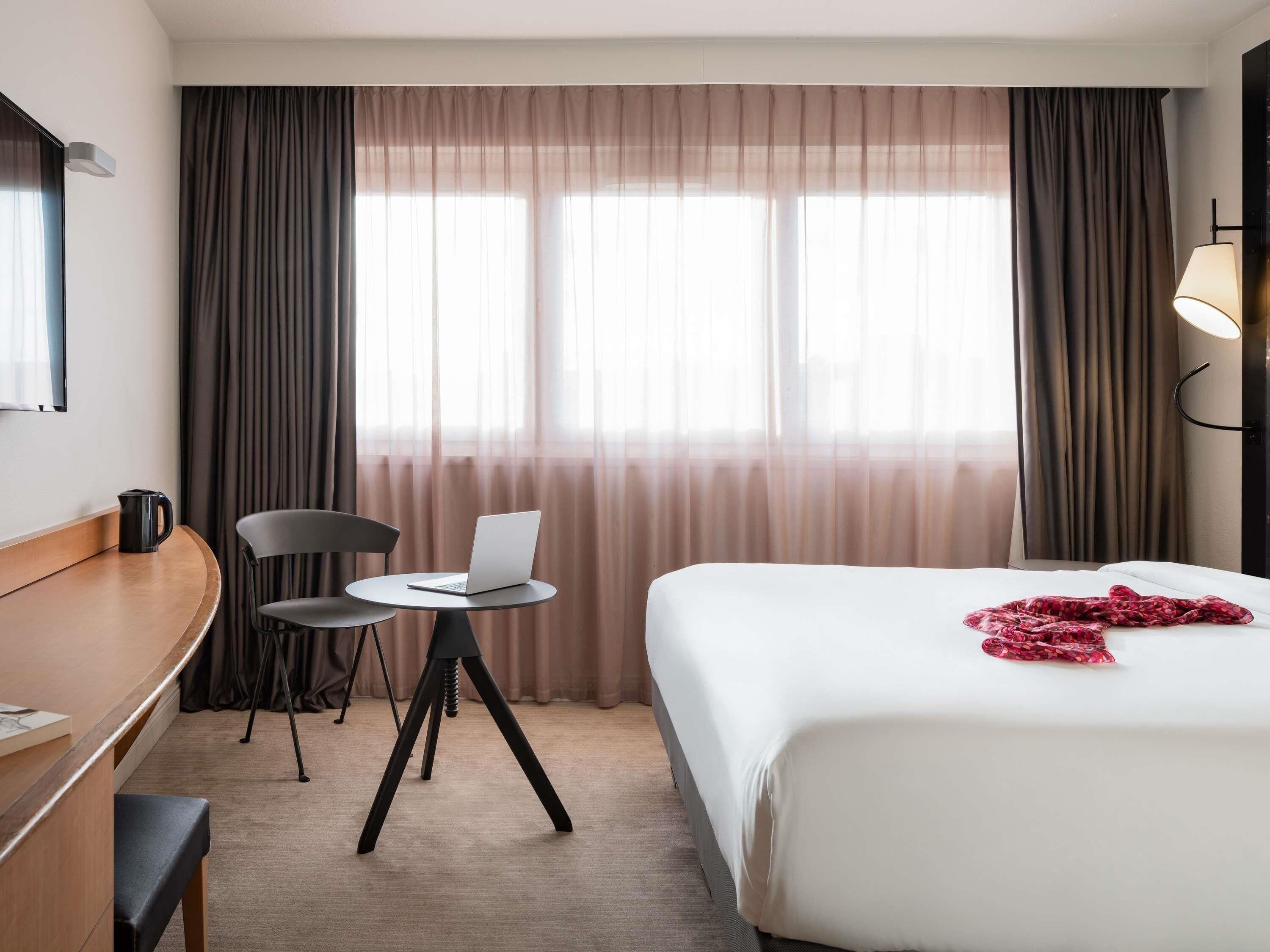 A hotel room with a bed, a desk with a laptop, a chair, and a window with curtains.