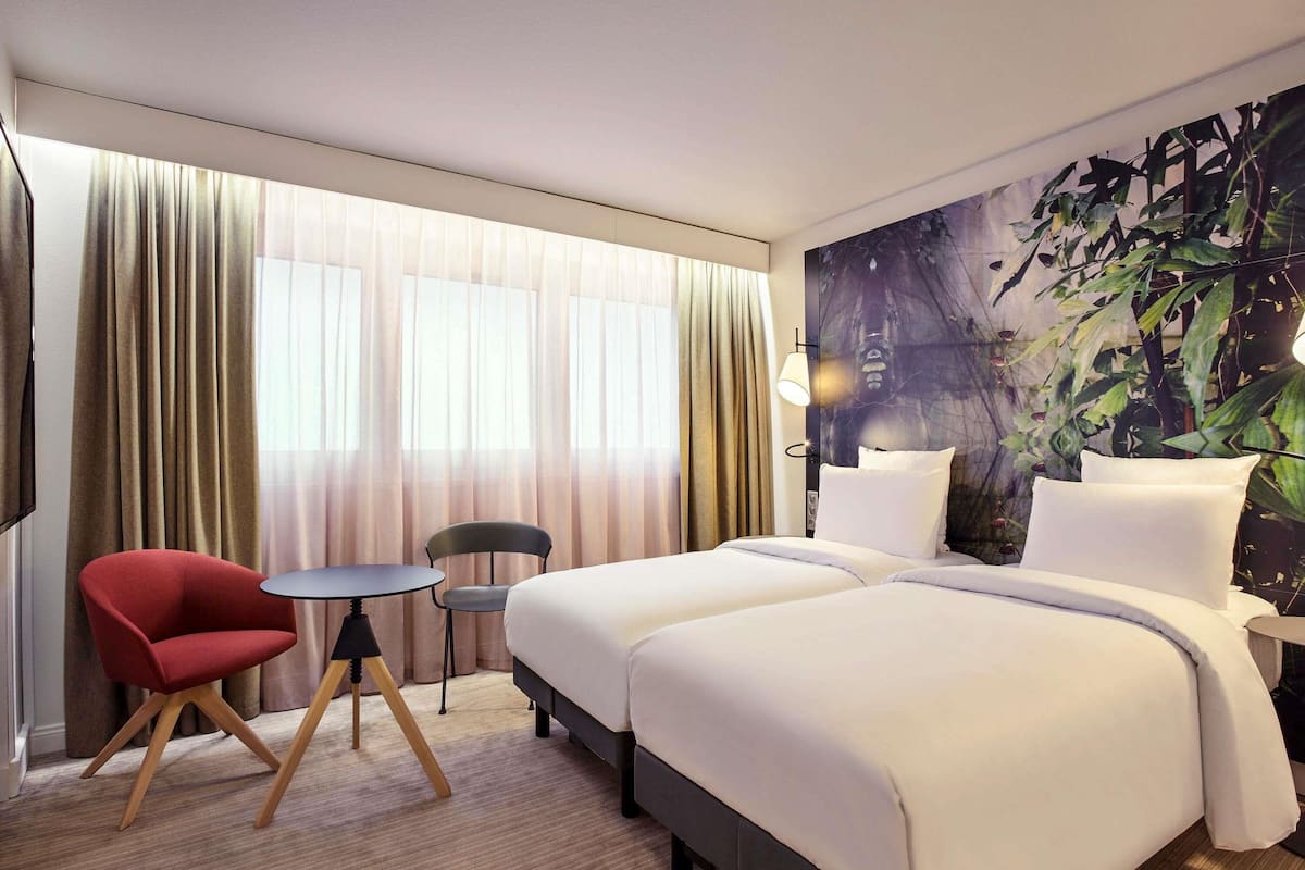 Ett hotellrum med en säng, två stolar, ett litet bord och ett stort fönster med gardiner.