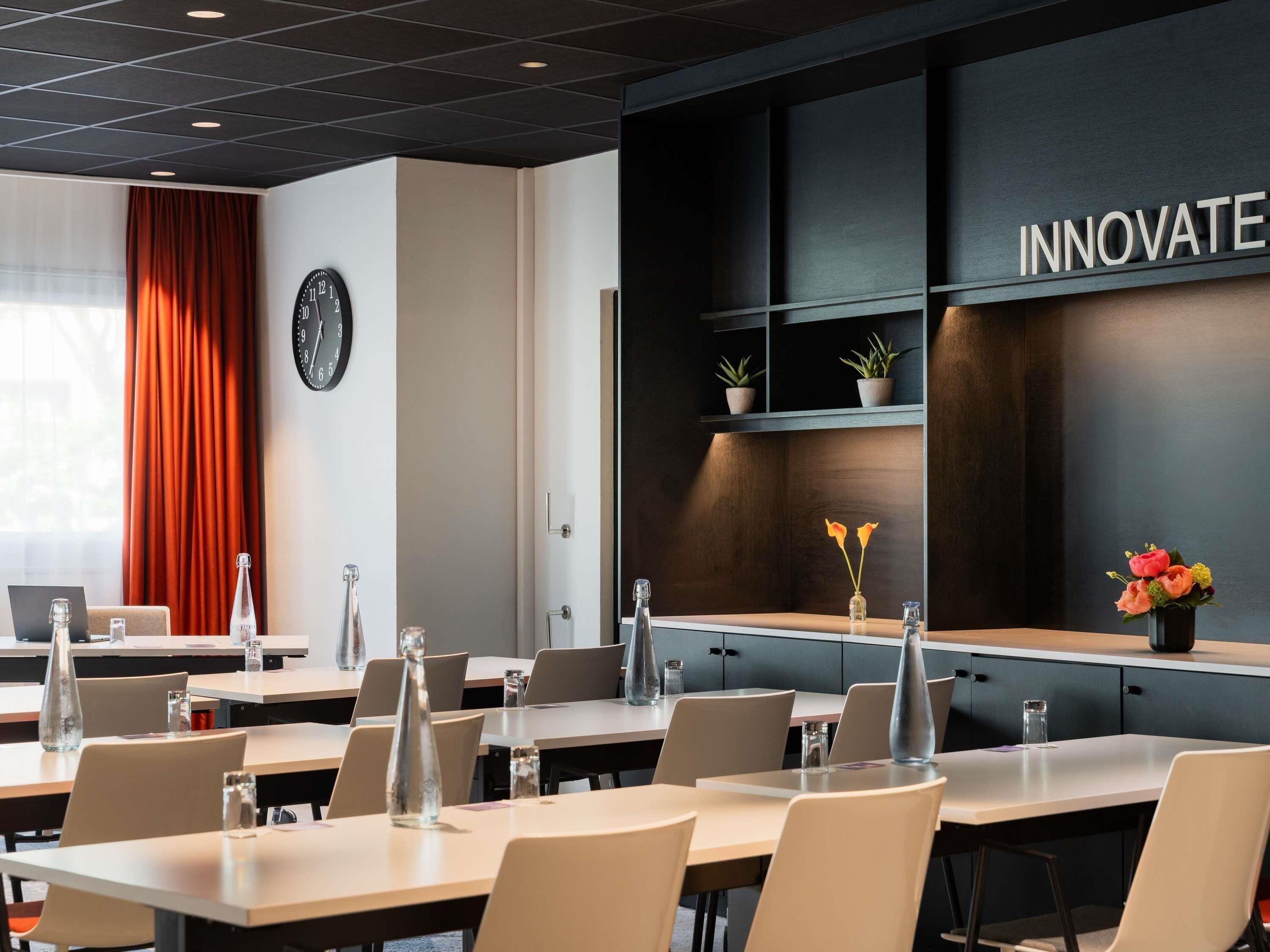 A modern conference room with tables, chairs, a clock, a shelf with plants, and a wall with the word 'INNOVATE'.