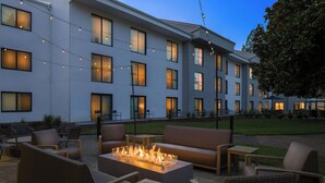 An outdoor seating area with wicker furniture and a fire feature, set against a building with multiple windows and string lights. - Courtyard by Marriott Portland Beaverton (Beaverton)