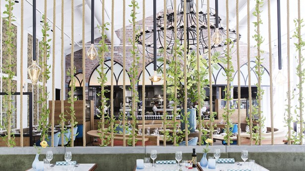 An indoor dining area with tables set for two, a chandelier, and a view of the kitchen through a glass partition. - Kasteel Kerckebosch (Zeist)