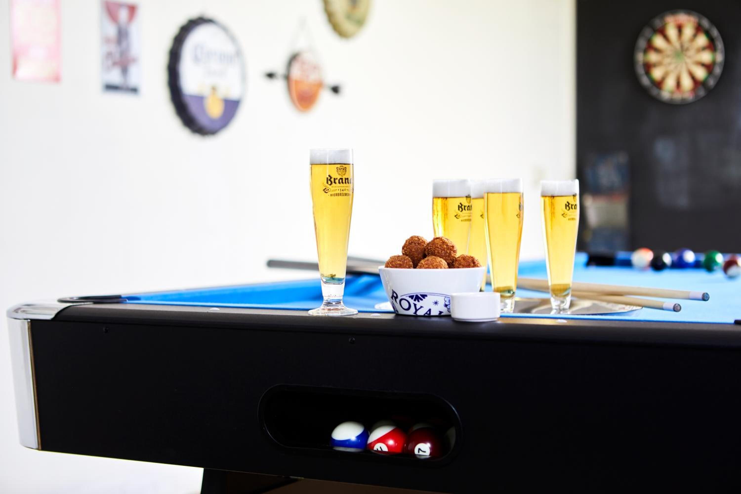 Una mesa de billar con cuatro vasos de cerveza, un bol de aperitivos y bolas de billar.