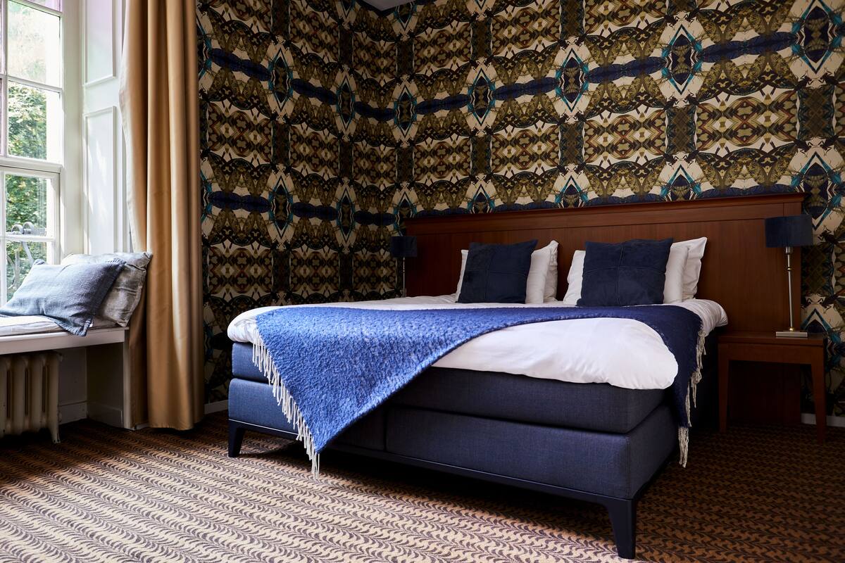 Un dormitorio con papel tapiz estampado, una cama con manta azul, una ventana con cortinas y una mesita de noche con lámpara.