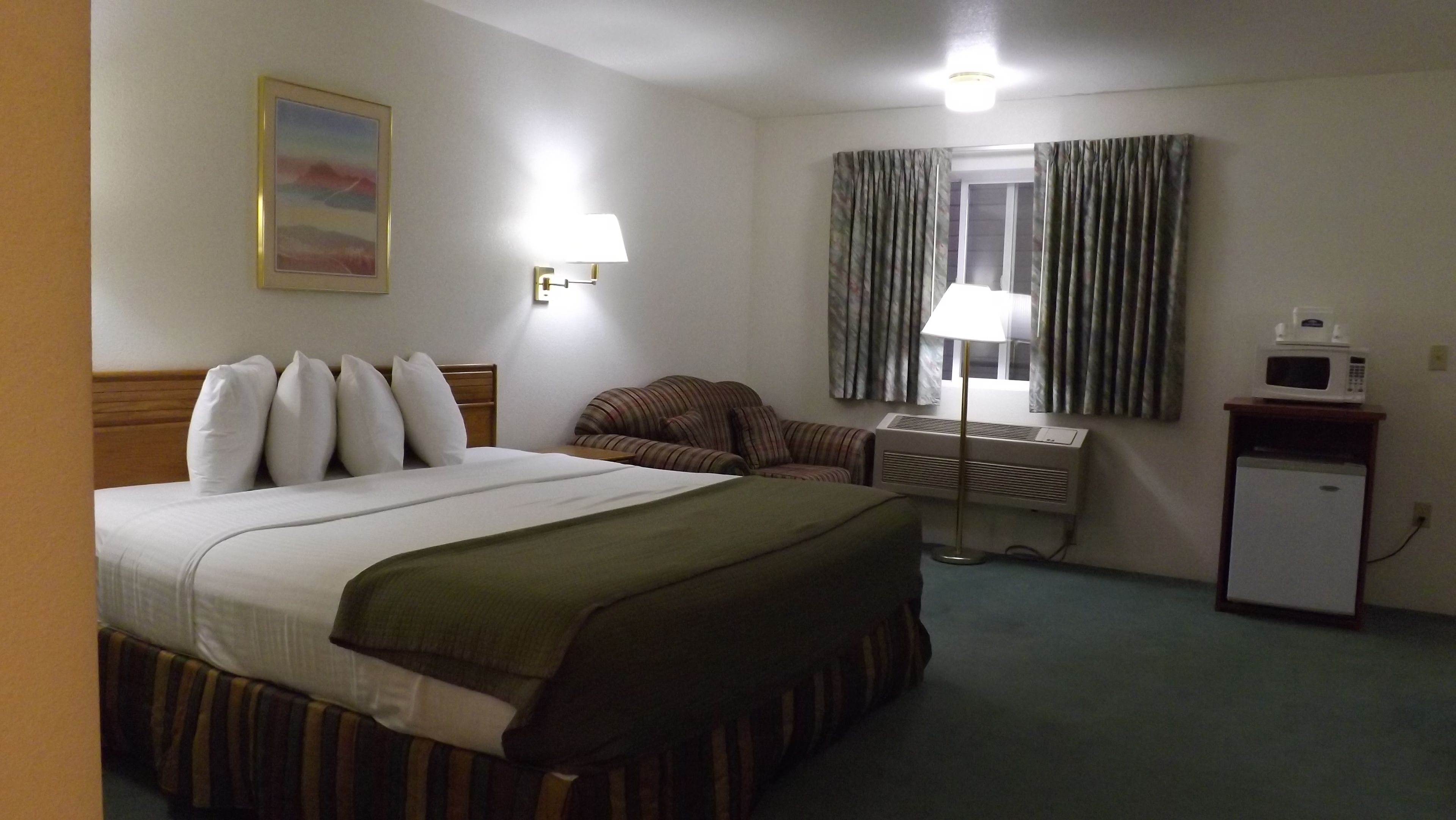 A hotel room with a bed, a chair, a desk, a lamp, a microwave, and a refrigerator.