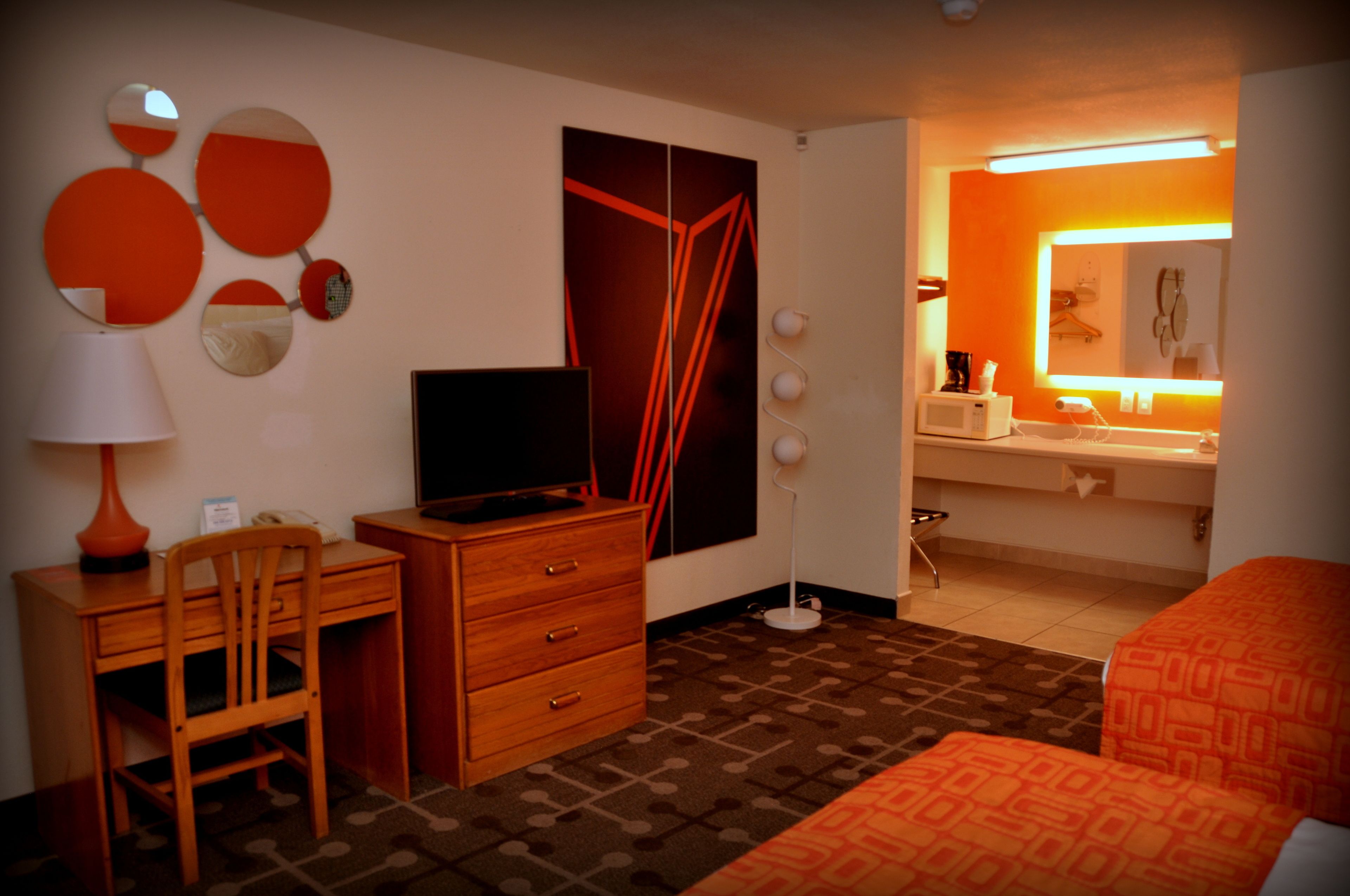 Een hotelkamer met een bureau, stoel, televisie, dressoir, kamerbreed tapijt met een patroon en een oranje sprei.