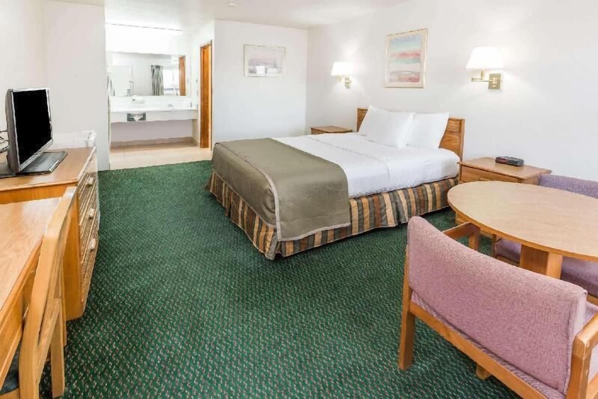 Habitación de hotel con cama, escritorio, silla, mesa redonda, televisor y baño con espejo, además de dos lámparas fijadas a la pared.