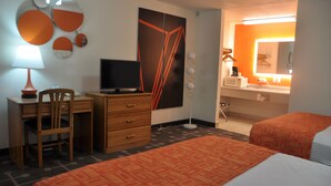 Habitación de hotel con escritorio, silla, televisión, cama y baño con espejo y lavabo.
