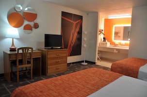 Quarto de hotel com escrivaninha, cadeira, televisão, cama e banheiro com espelho e pia.