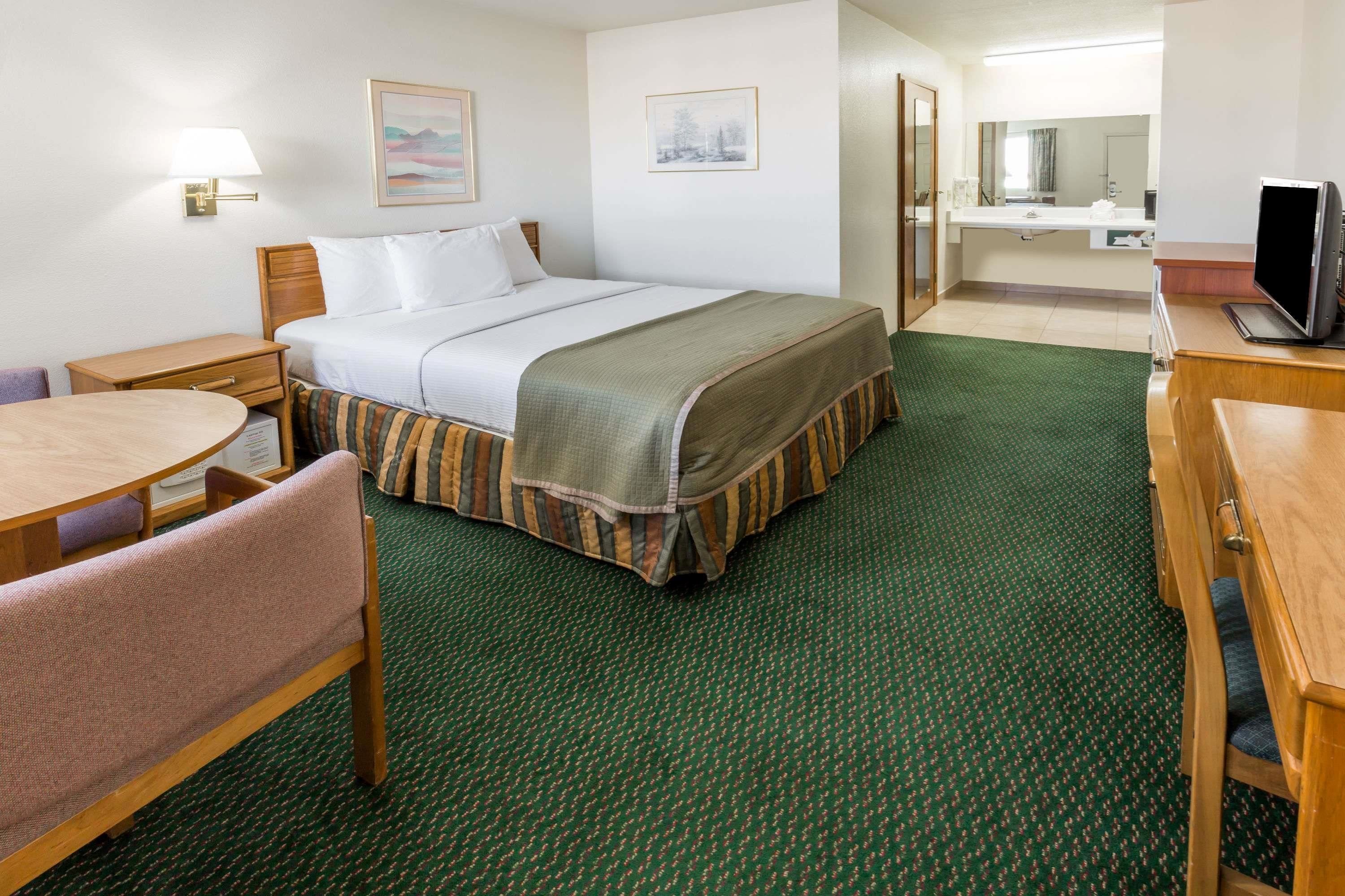 Habitación de hotel con cama, escritorio, silla, pequeña mesa, lámpara, televisor y baño con espejo.
