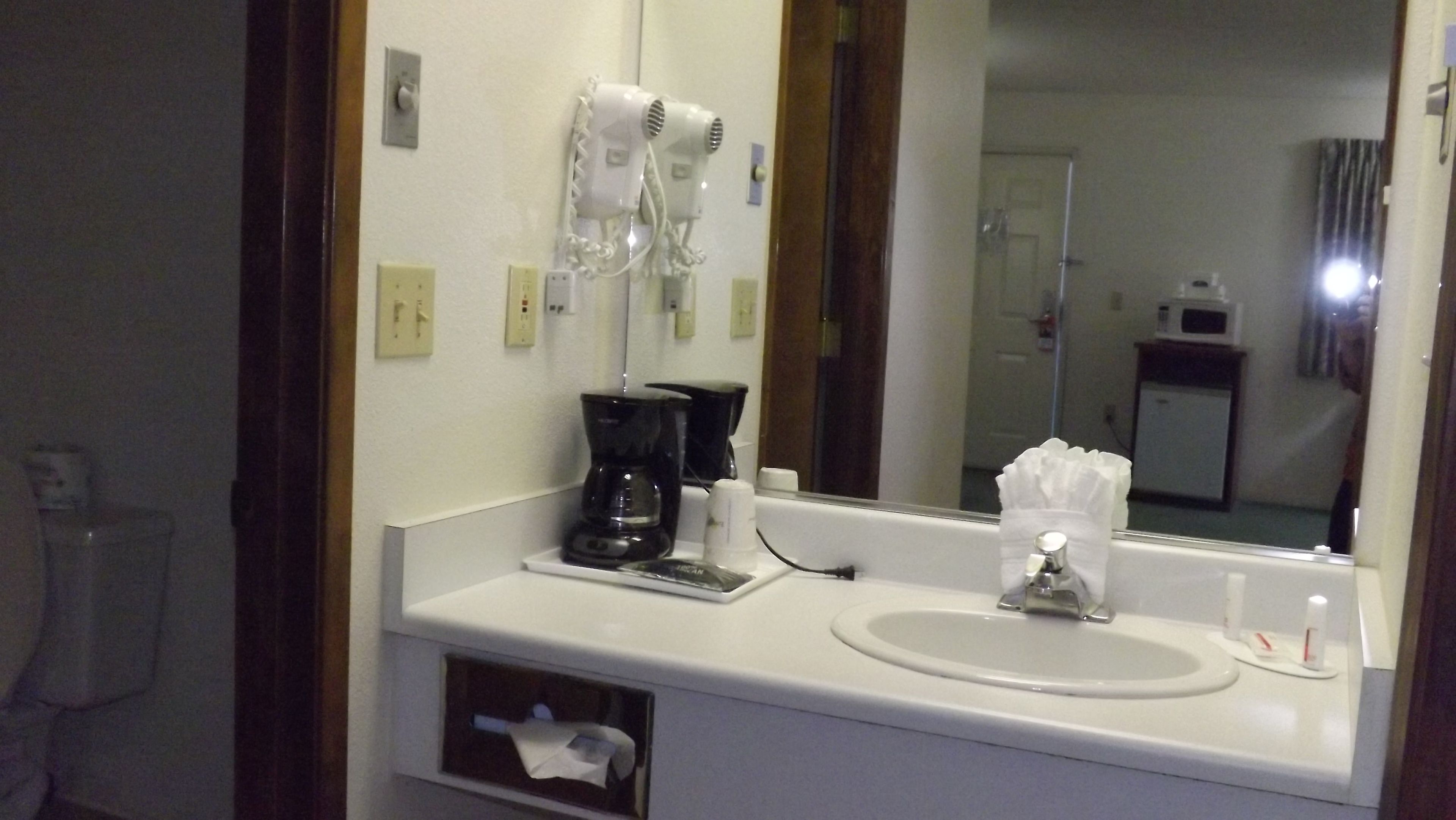 Une salle de bain d’hôtel avec un lavabo blanc, un miroir, un sèche-cheveux, une machine à café et un distributeur de mouchoirs.