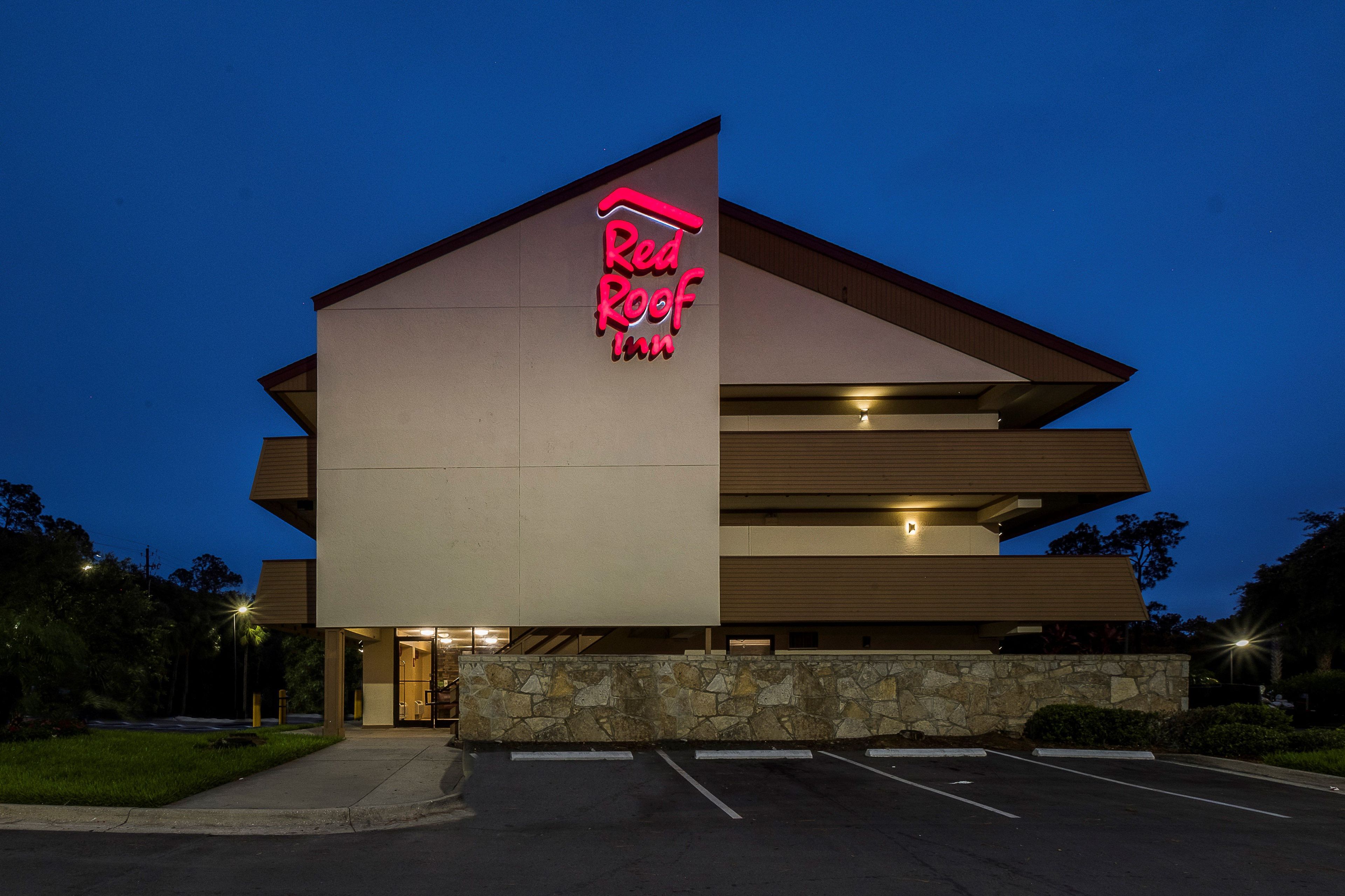 A Red Roof Inn with a modern architectural design, featuring a prominent neon sign, a parking lot, and a stone facade.