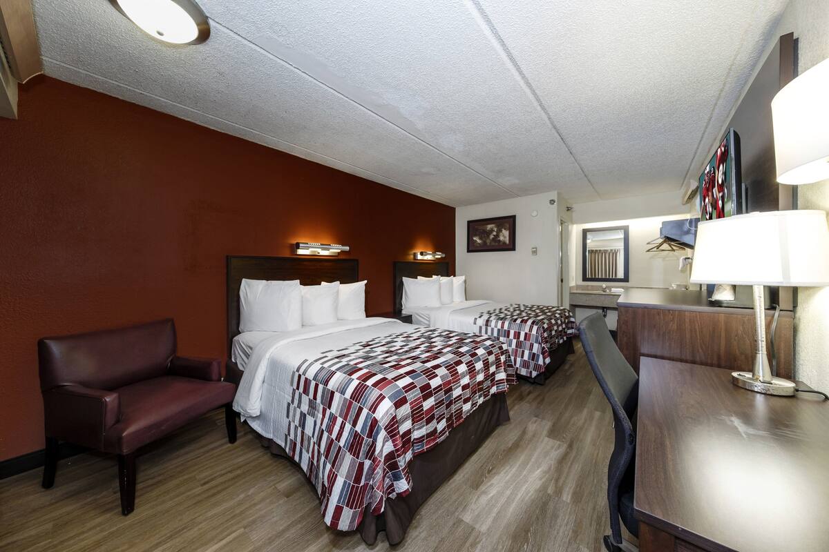 A hotel room with two beds, a desk, a chair, and a lamp.
