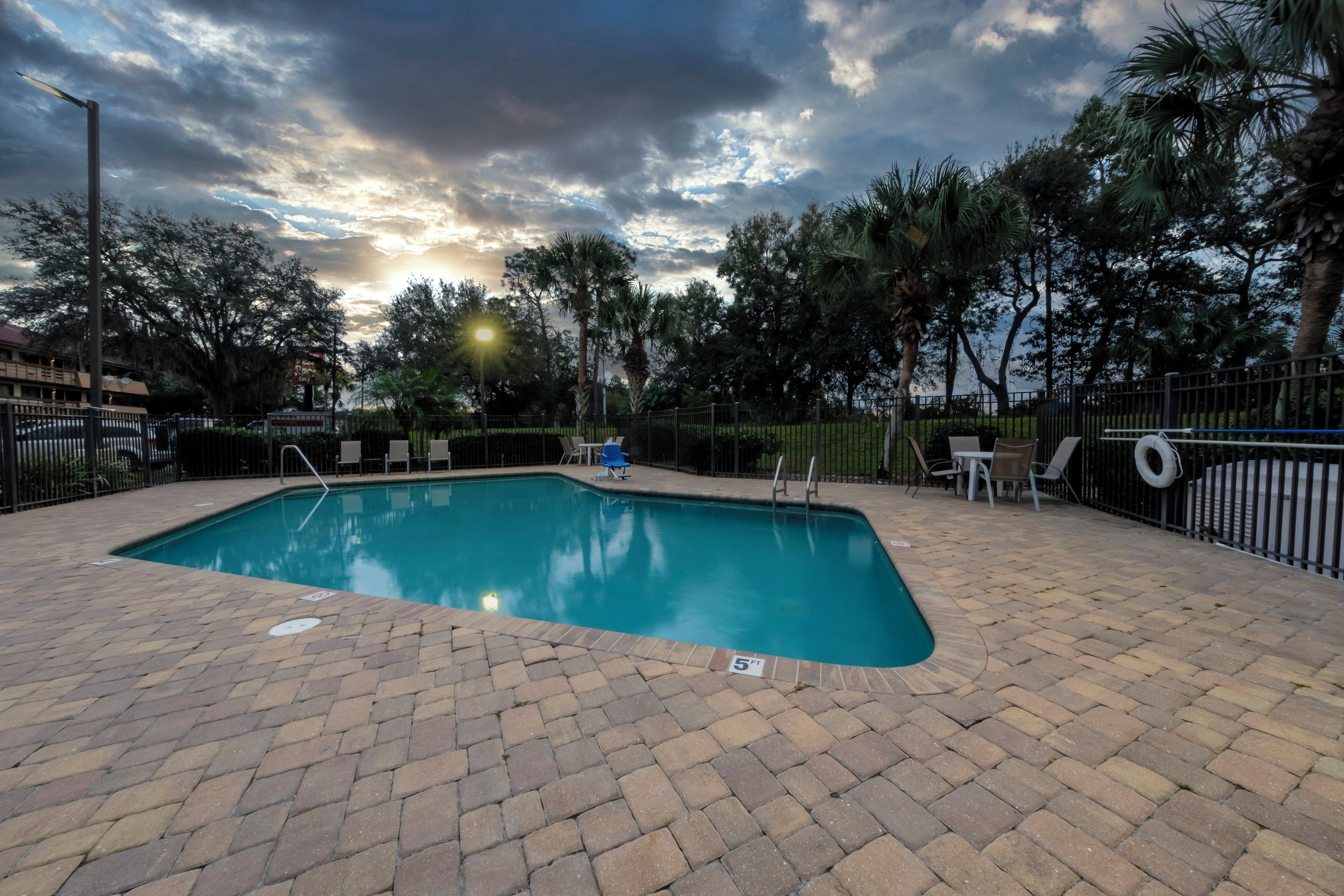 Une piscine rectangulaire bordée d’une terrasse en briques, entourée d’une clôture et d’arbres.