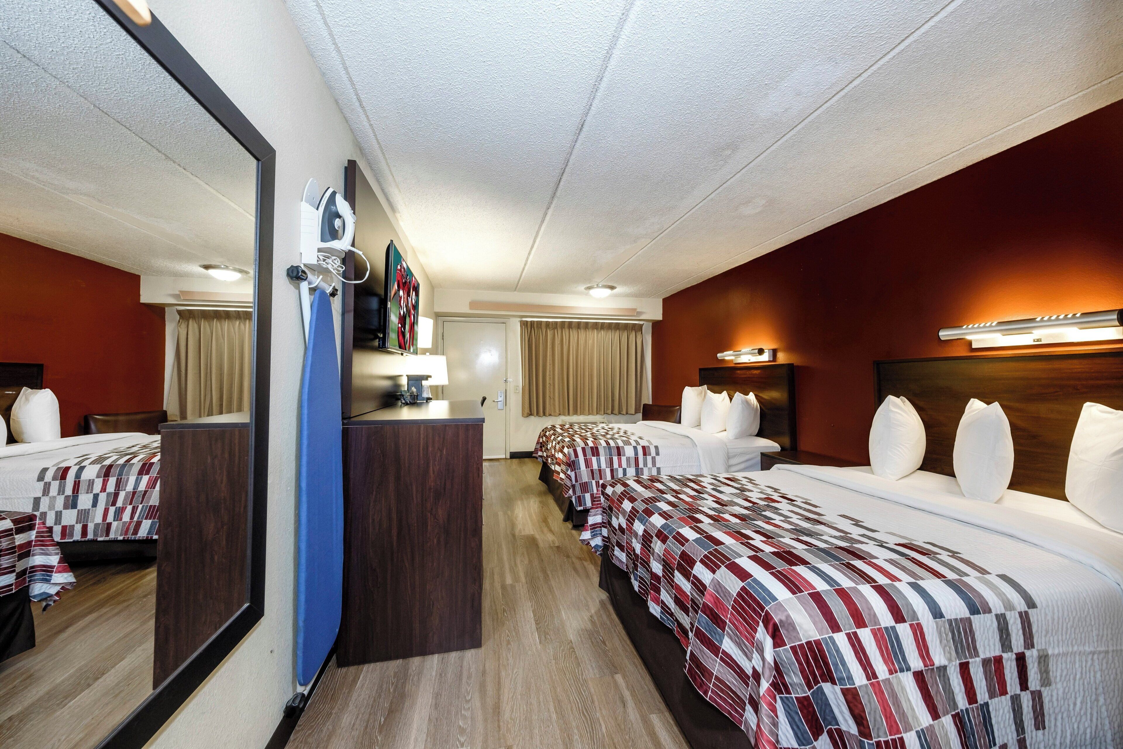 A hotel room with a mirrored wall, a desk with a lamp, a bed with a checkered bedspread, and a headboard with a light fixture.