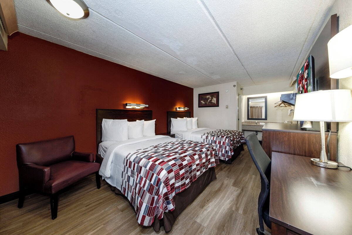 A hotel room with two beds, a desk, a chair, a mirror, and a wall-mounted TV.