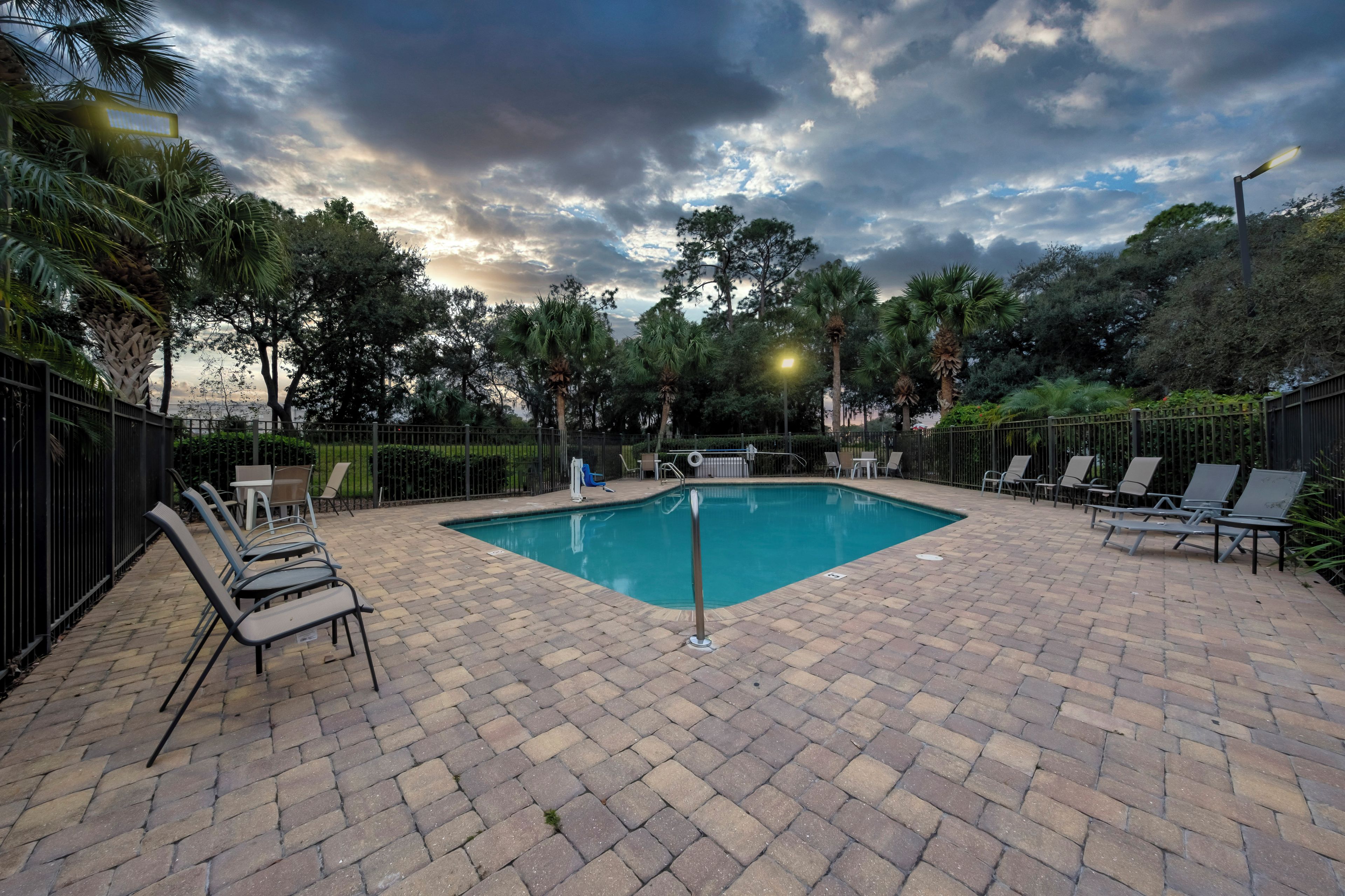 A rectangular pool with a surrounding paved area, equipped with lounge chairs and a fence.