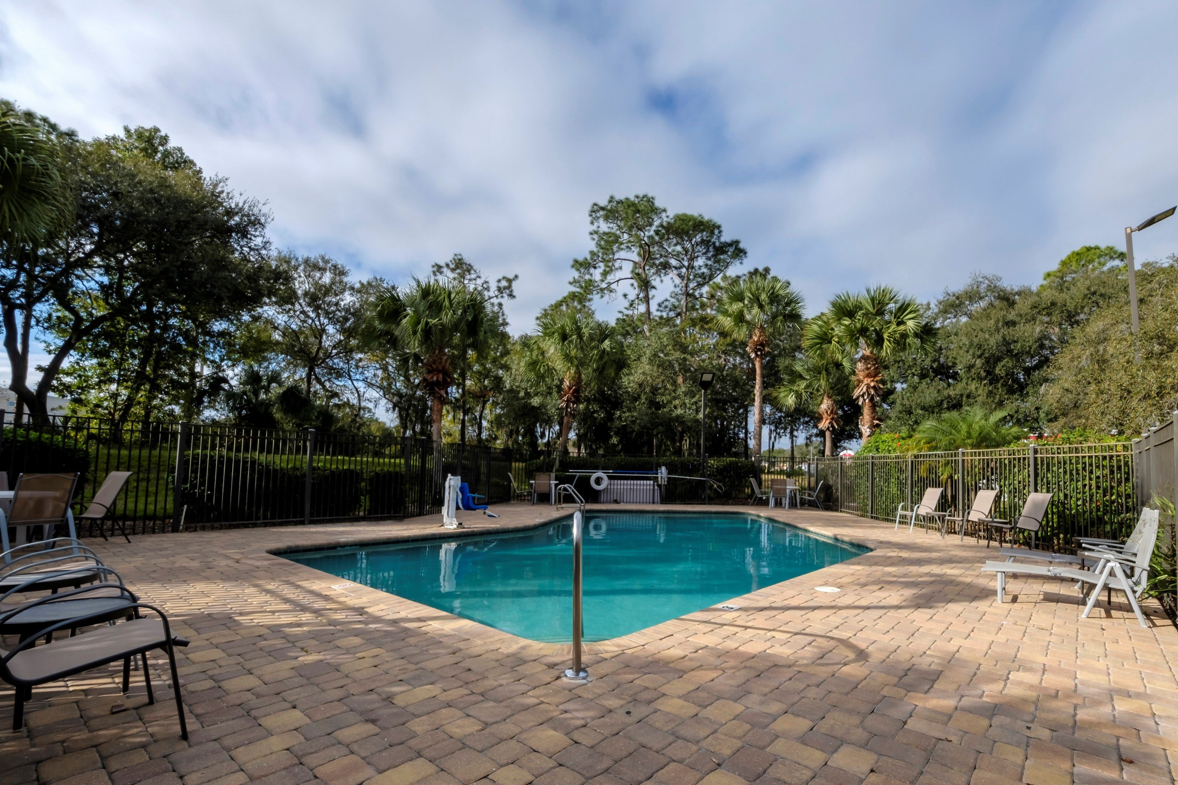 Una piscina rectangular con un patio de ladrillo, rodeada de sillas de descanso y una valla de metal.