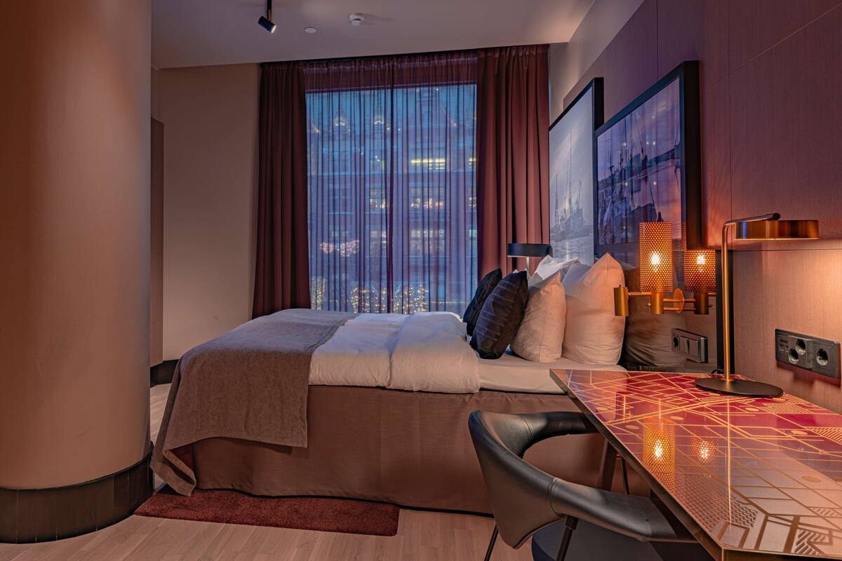 A modern hotel room with a bed, a desk, and a view of the cityscape through large windows.