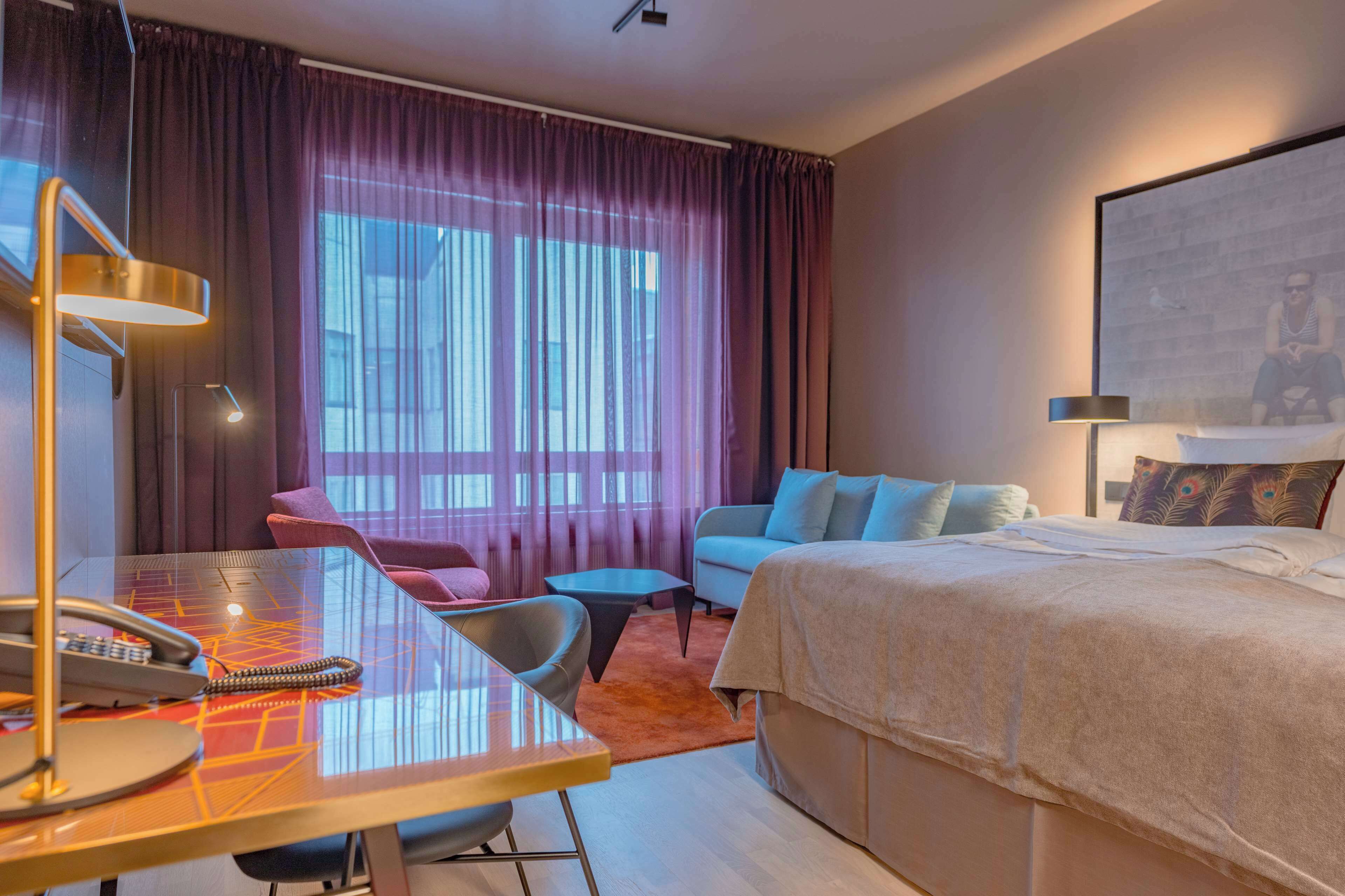 Une chambre d’hôtel comprenant un lit, un bureau avec un téléphone, une chaise, un canapé et une grande fenêtre avec des rideaux.