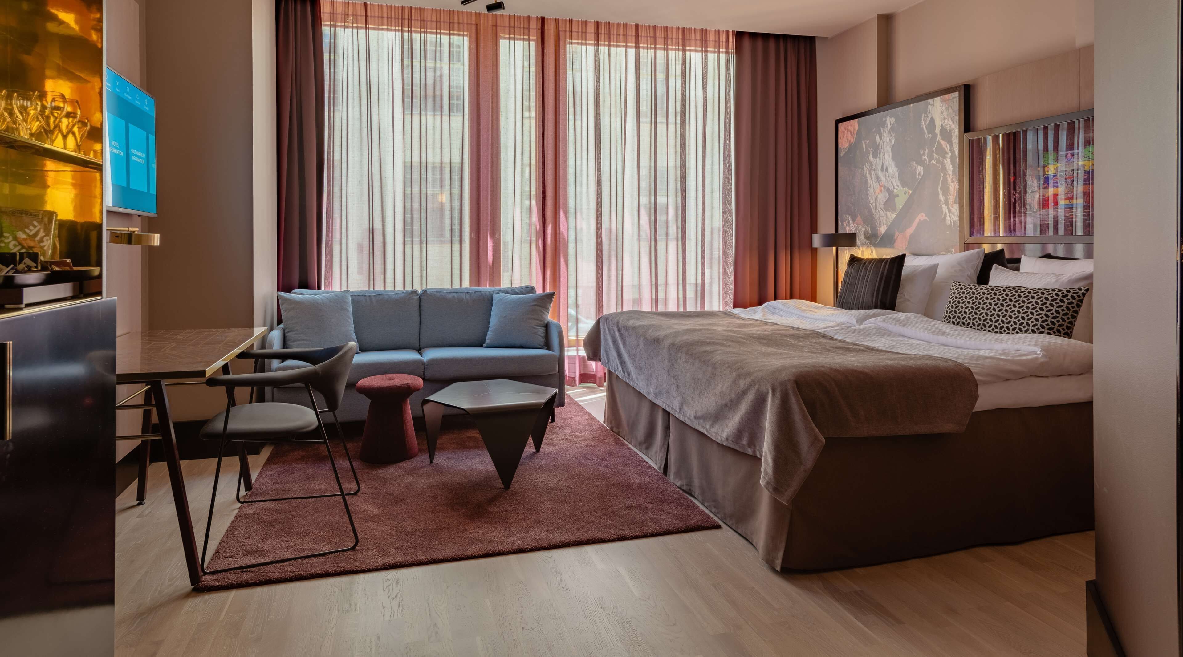 A modern hotel room with a sofa, a small table, a bed, and a TV.