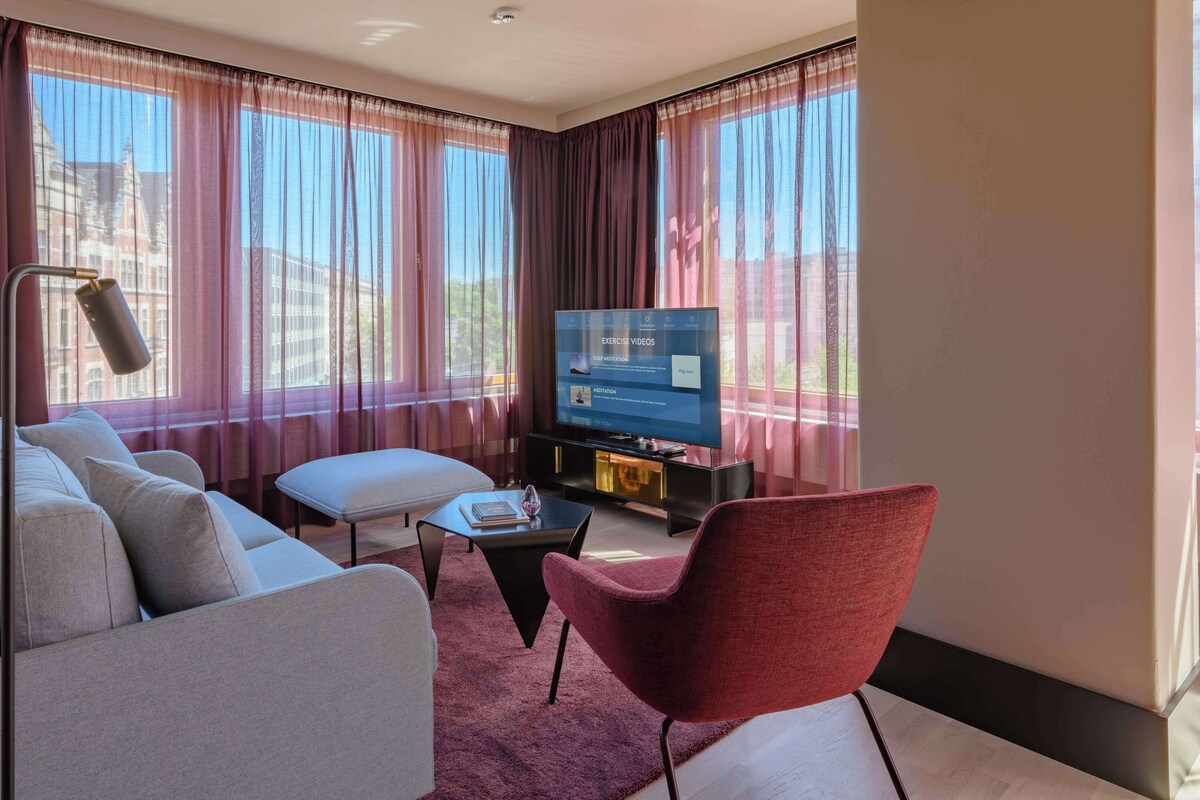 Una sala moderna con un sofá, un sillón y un reposapiés, un televisor de pantalla plana y ventanas amplias con cortinas translúcidas.