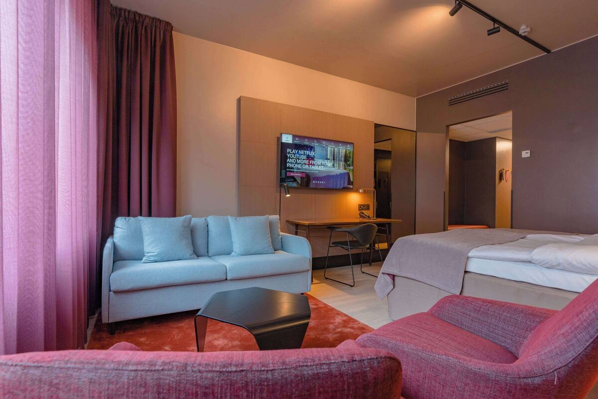 A hotel room with a light blue sofa, a pink armchair, a small black table, and a bed with a grey blanket.